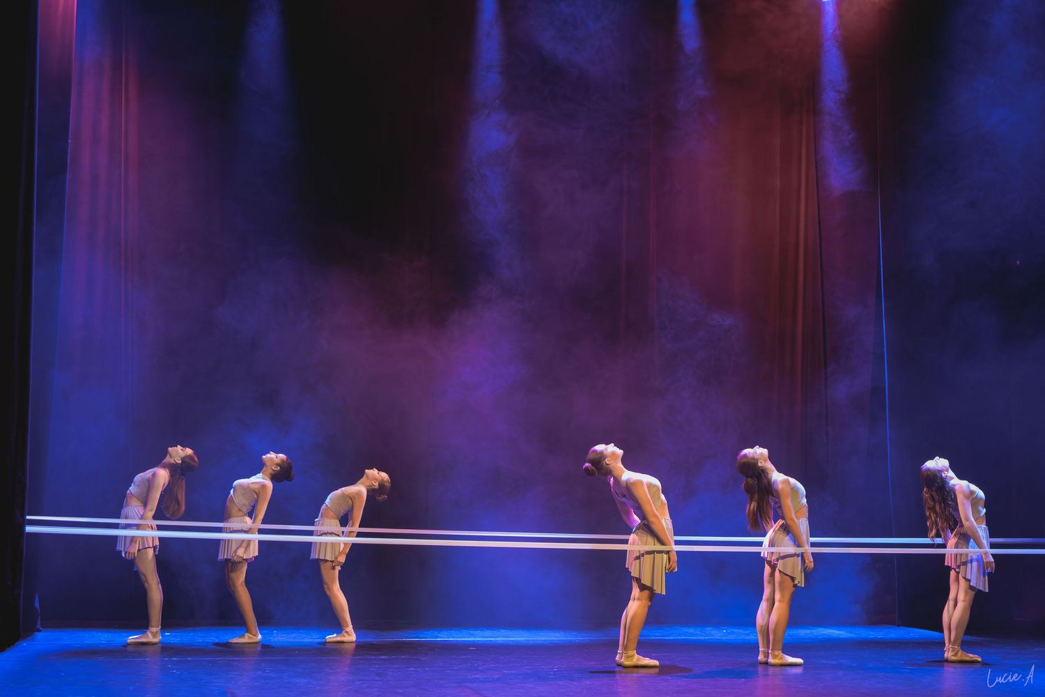 Six danseurs saluent à l'unisson sur une scène plongée dans le brouillard, illuminée par un éclairage dramatique violet et bleu.