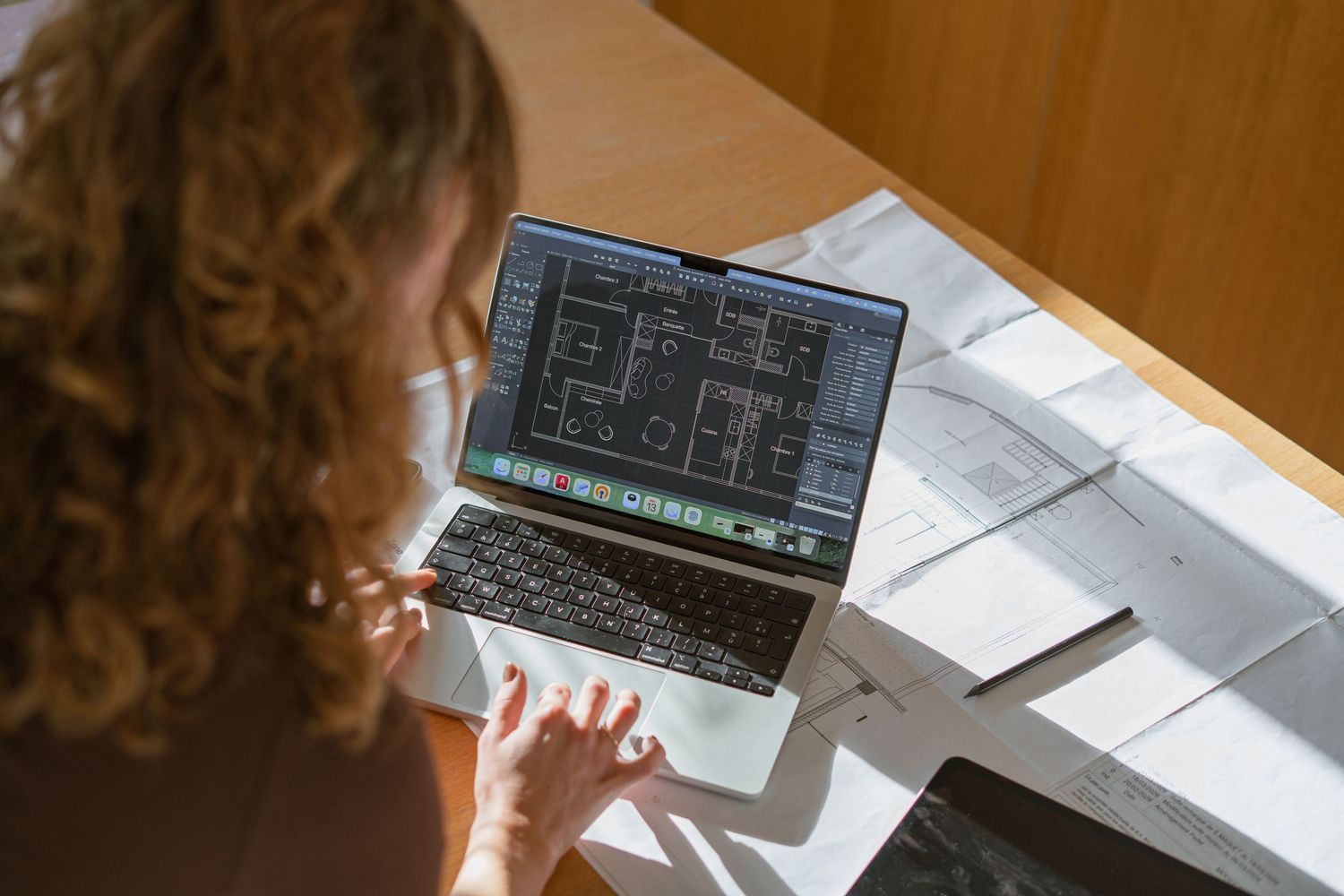 Woman with curly hair reviewing architectural floor plan drawings on a laptop at a desk covered with printed blueprints.