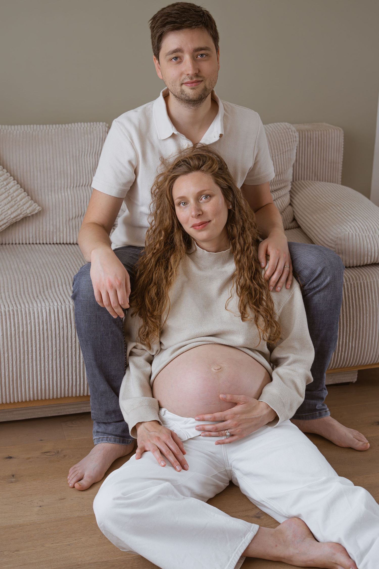 Pregnant woman sitting on floor leaning against partner near couch, both smiling calmly in neutral home setting.