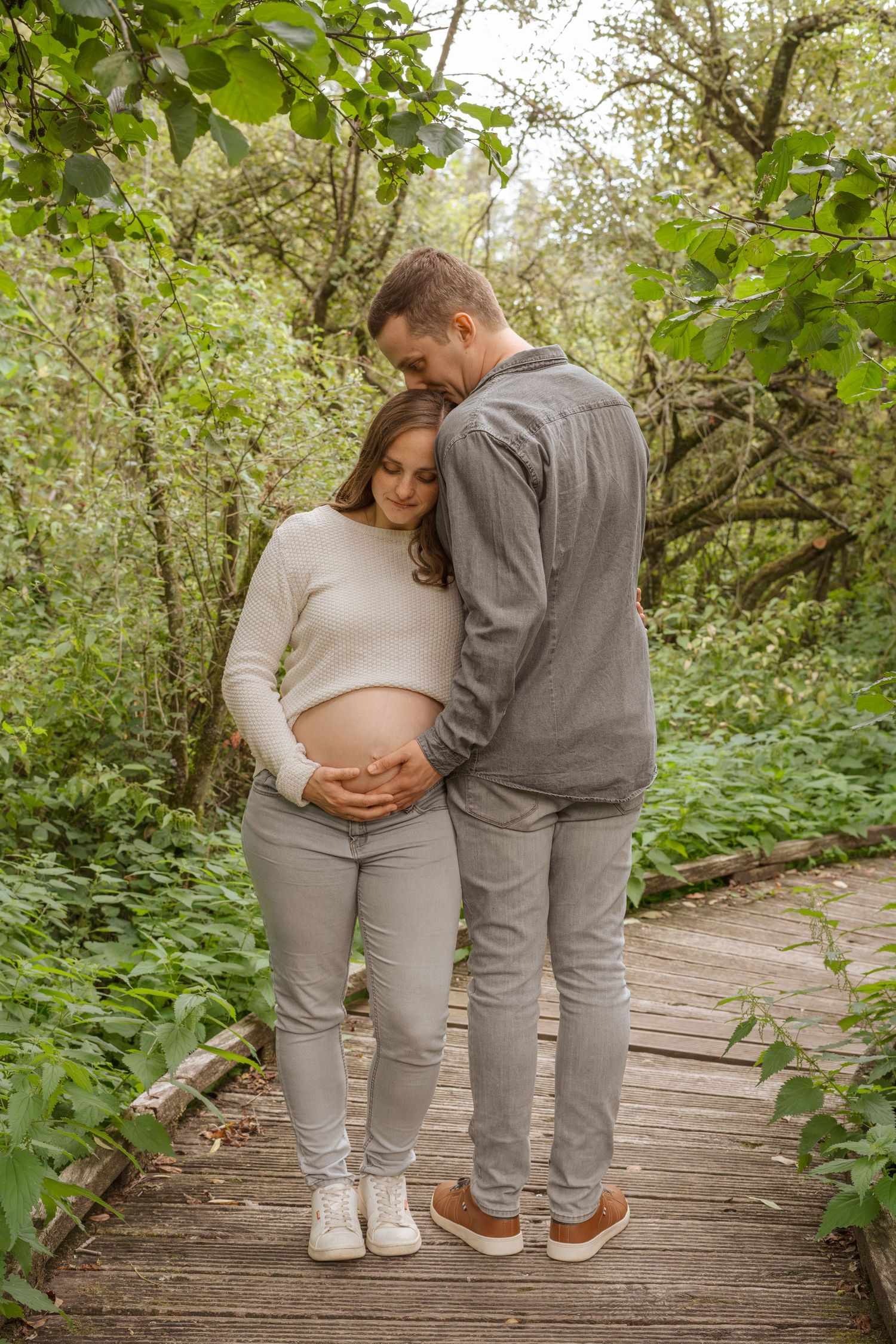 Pregnant couple embracing on wooden path in lush green forest, man tenderly holding woman's baby bump outdoors.
