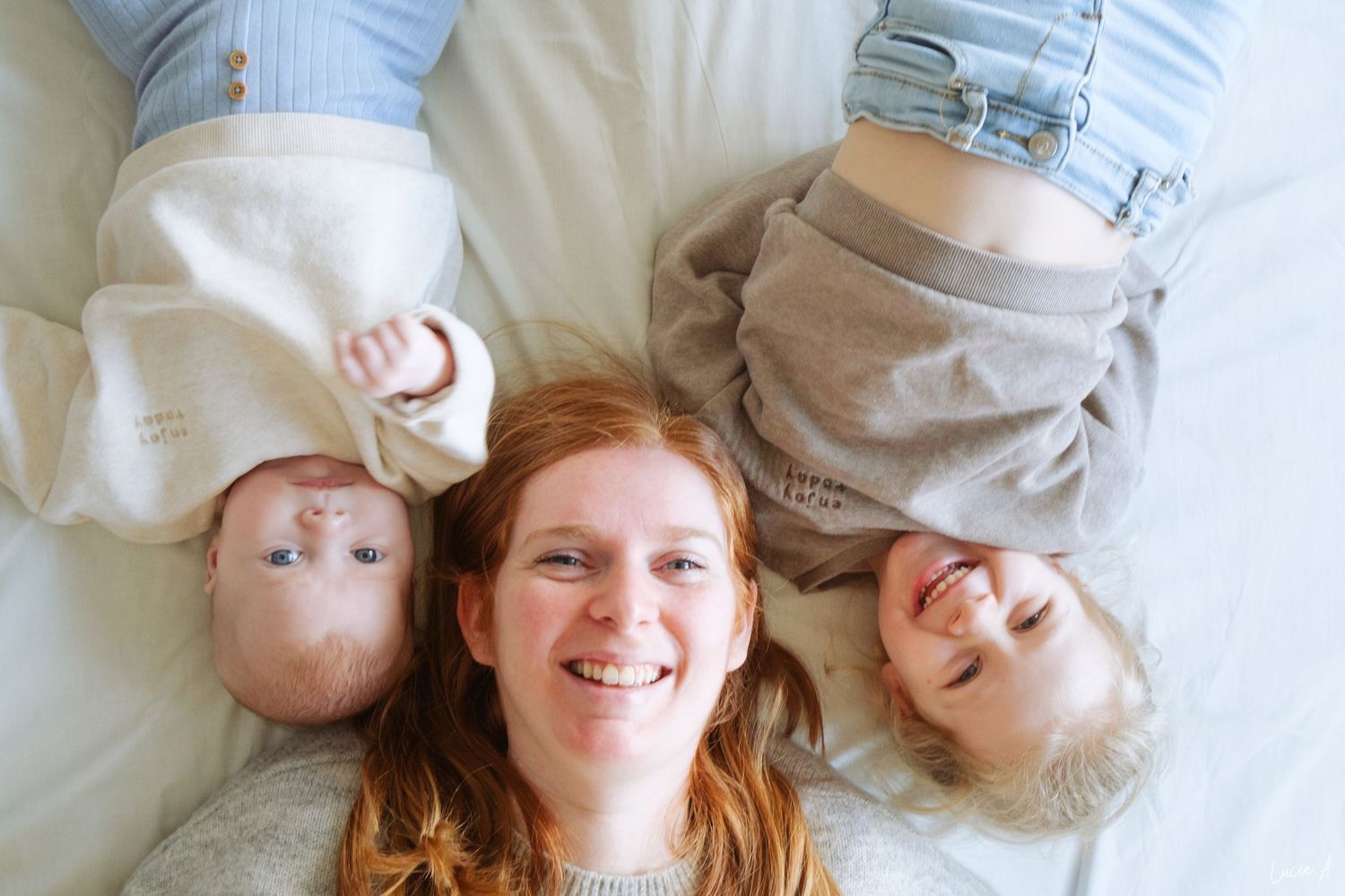 Portrait d'un famille réunissant une maman et ses deux enfants allongés sur un lit, tous souriants à la lumière naturelle