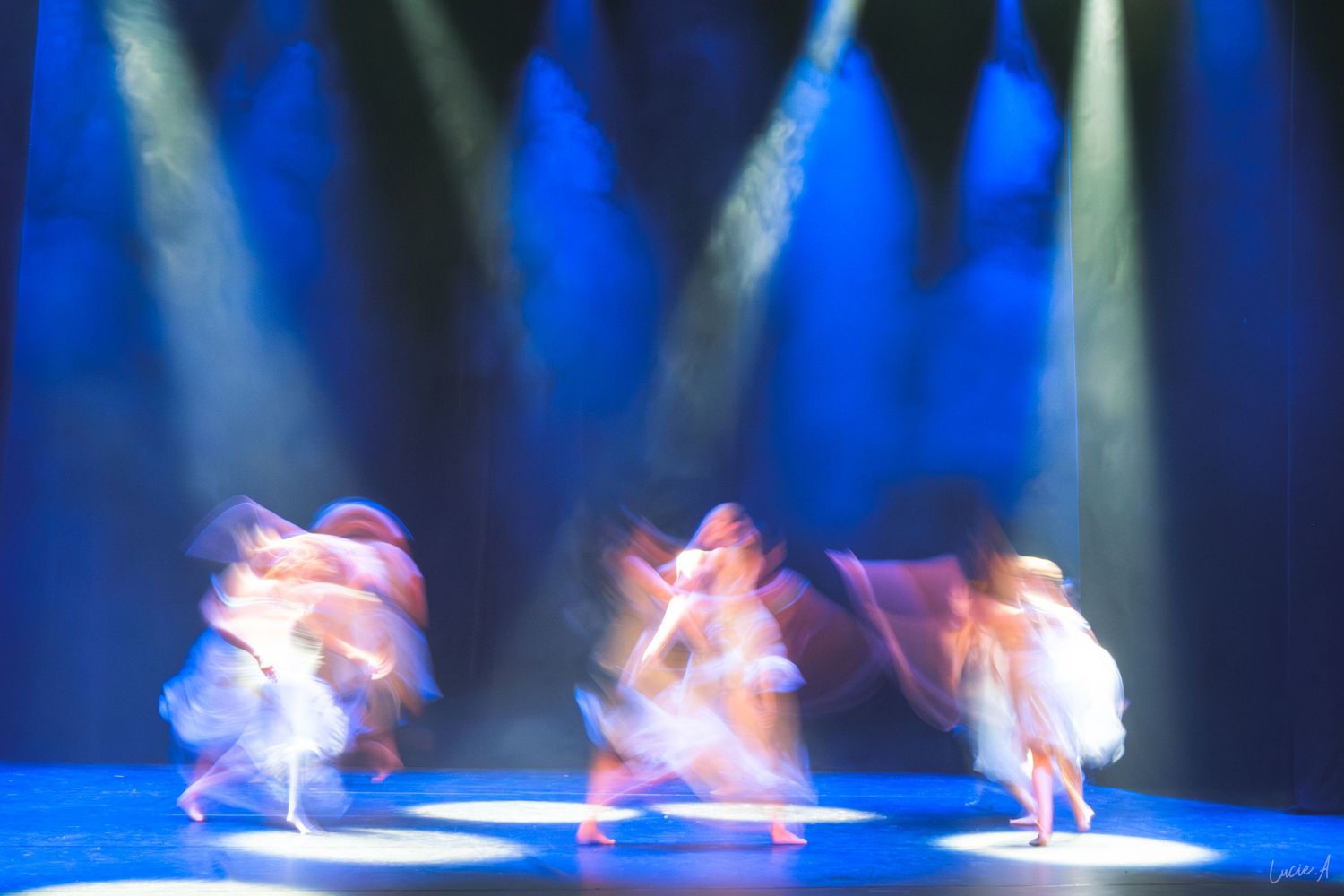 Mouvements flous de danseurs en costumes blancs évoluant sur une scène éclairée en bleu par des projecteurs spectaculaires.