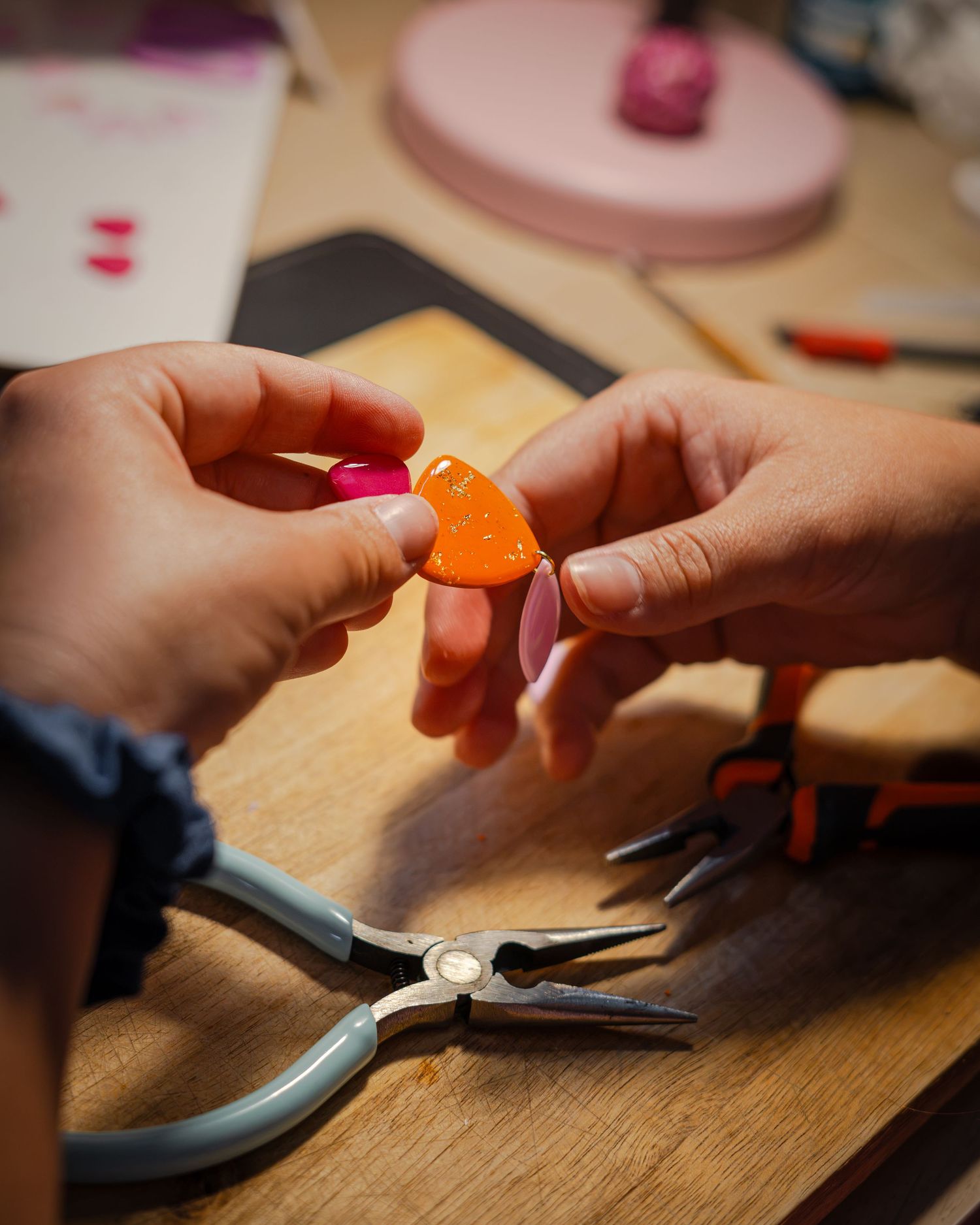Hands assembling orange and pink clay earrings with pliers on a wooden craft table.