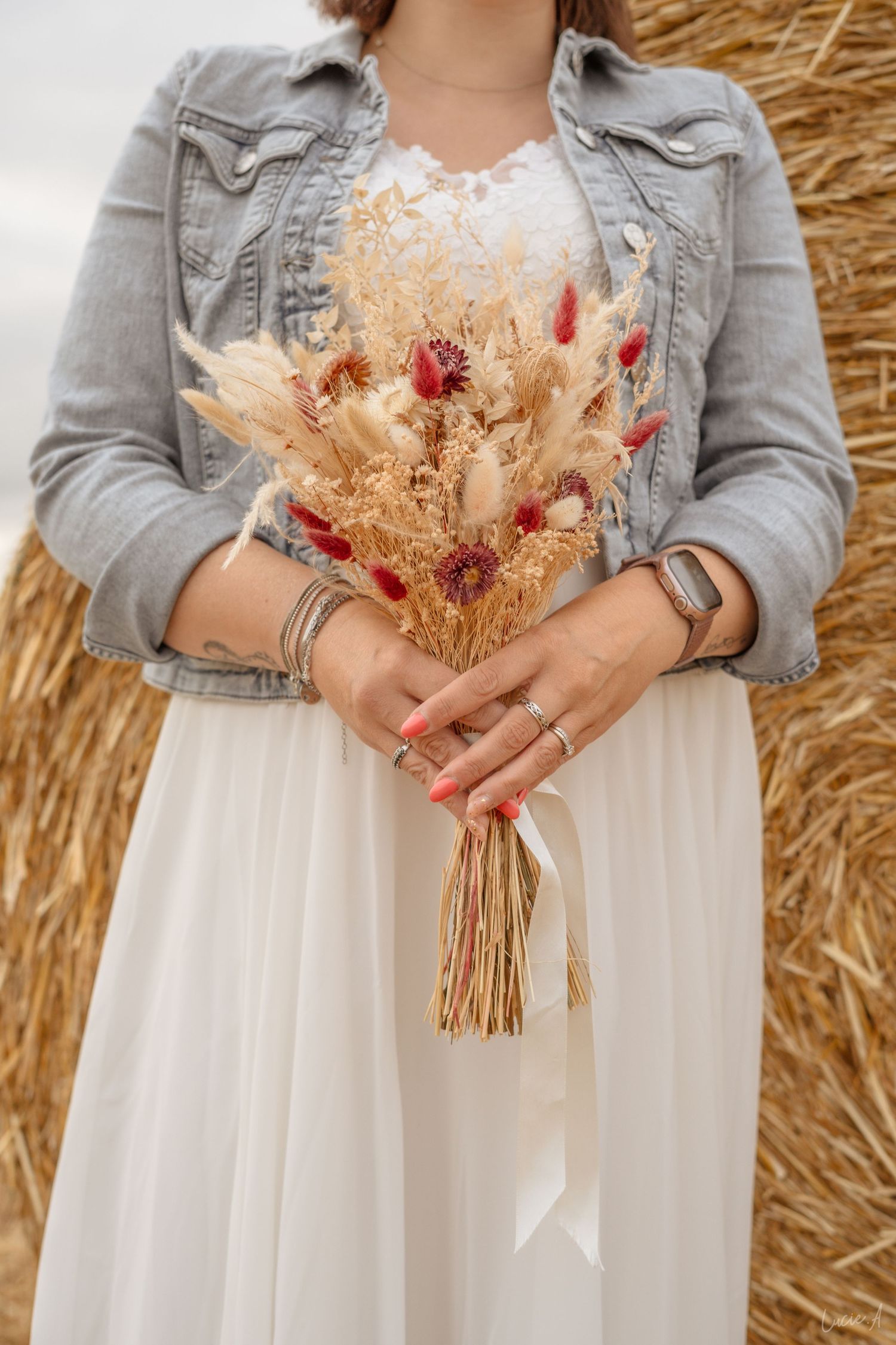 Femme en robe blanche et veste en jean tenant un bouquet rustique de fleurs séchées avec des touches de rouge près d'une botte de foin.