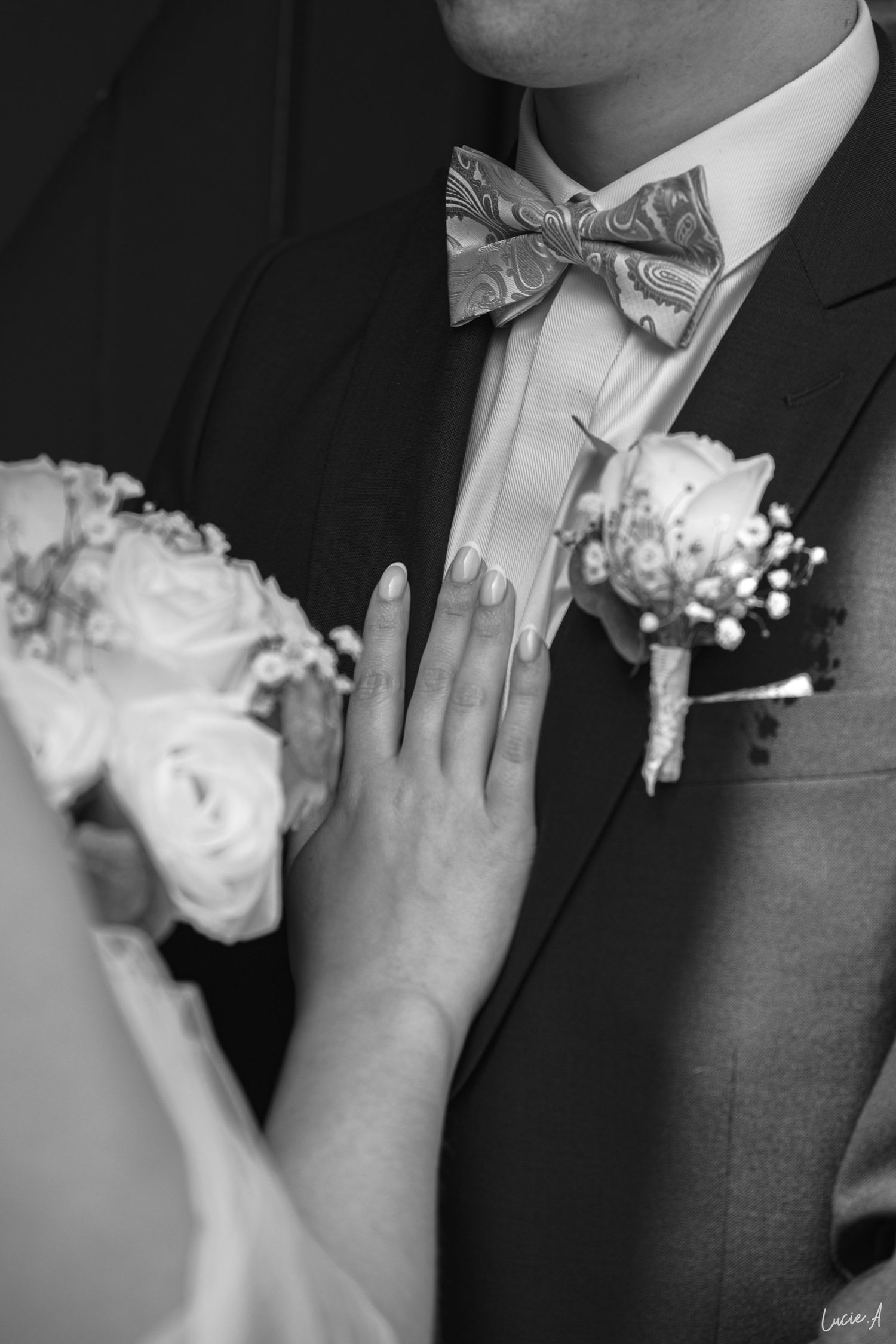 Photo en noir et blanc de la main de la mariée sur le costume du marié, avec boutonnière et bouquet.