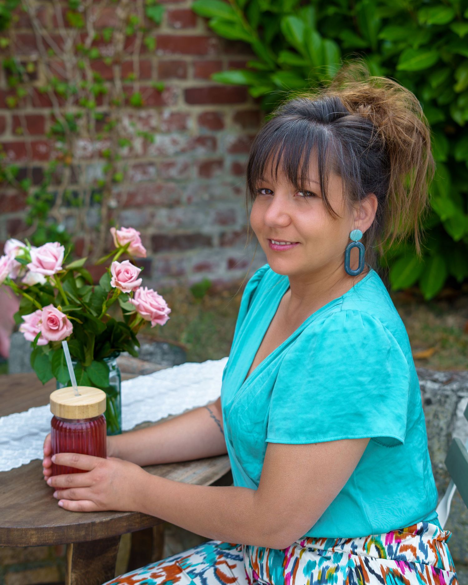 Woman in teal top holding a mason jar drink beside pink roses at an outdoor garden table.