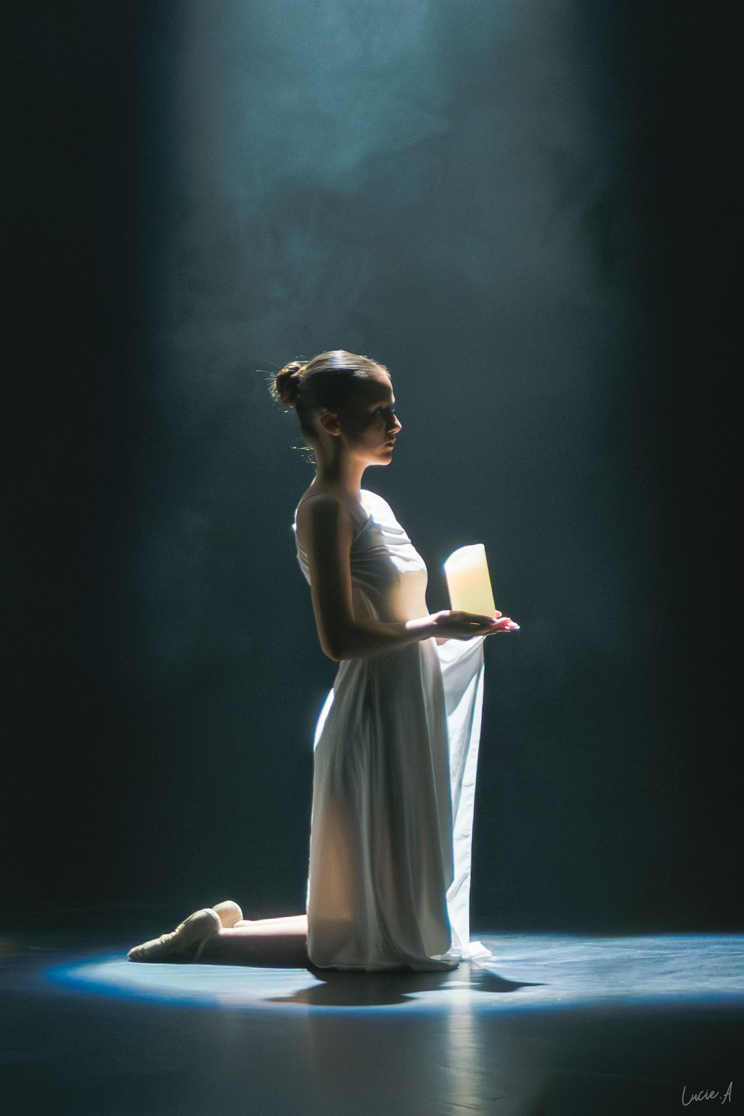 Une danseuse en robe blanche est agenouillée sur scène, tenant un objet lumineux sous un projecteur dramatique avec des effets de fumée.