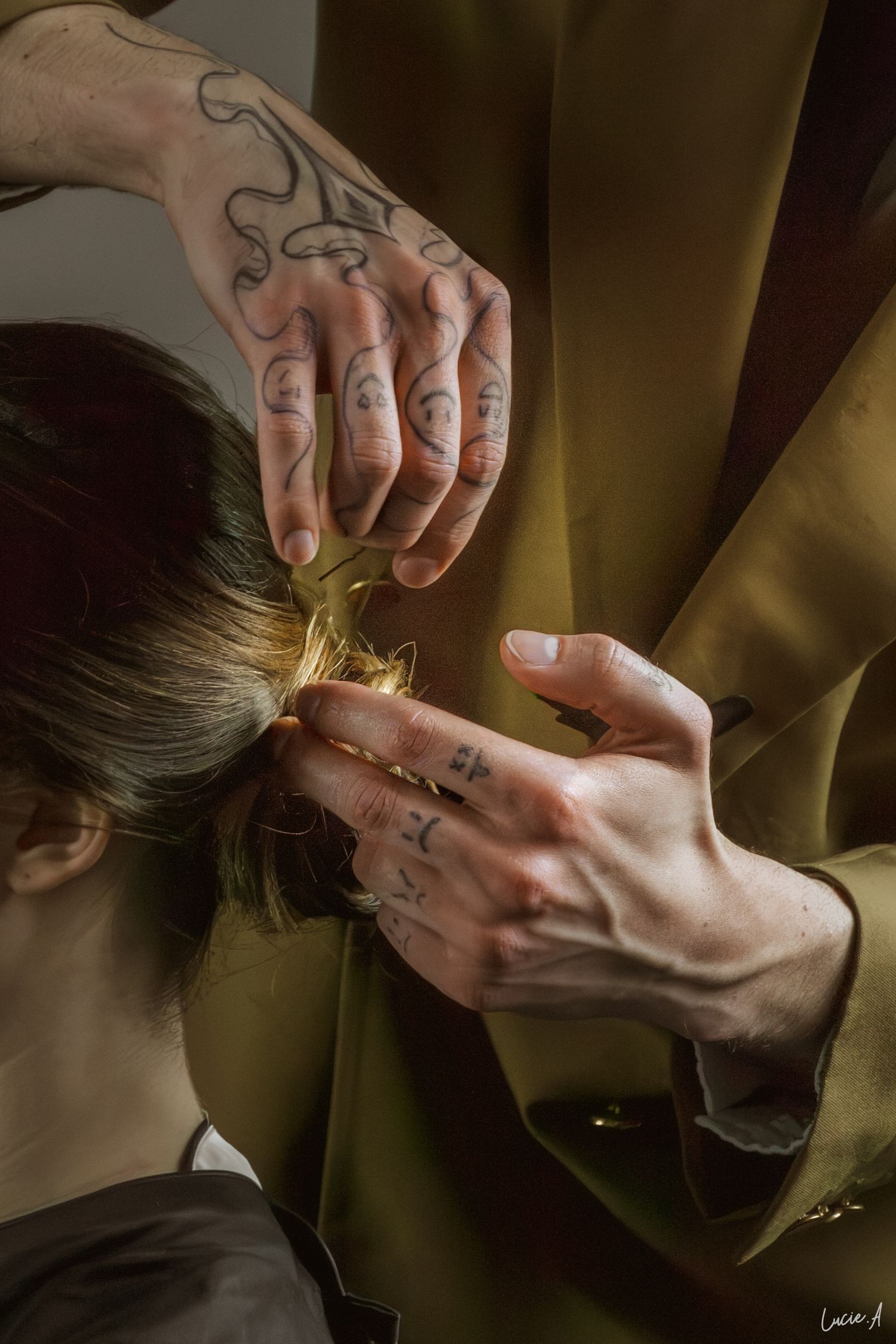 Des mains tatouées coiffent les cheveux d'une mariée en un chignon élégant sur un fond vert olive.