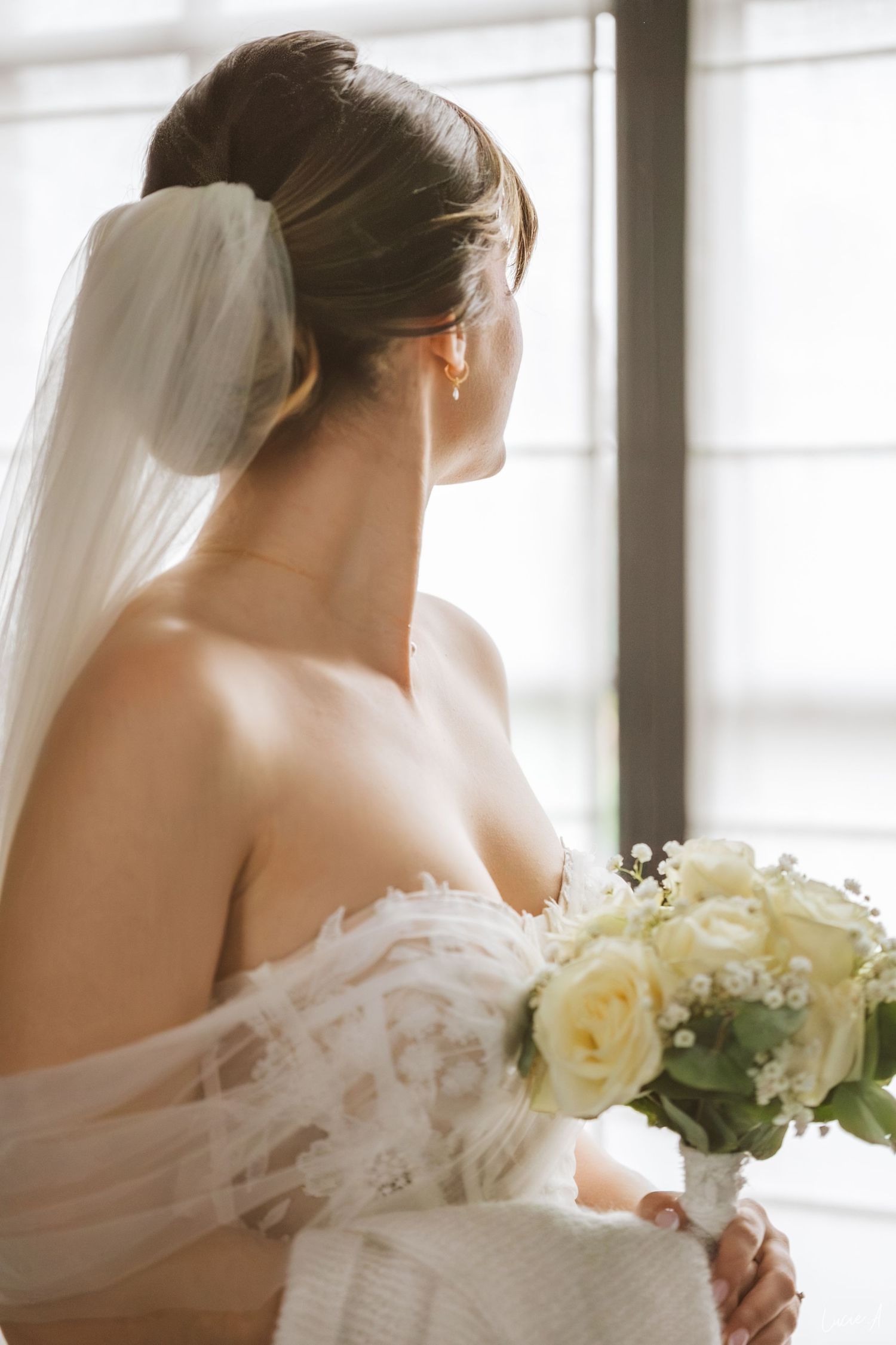 Élégante mariée en robe de dentelle bustier tenant un bouquet de roses blanches, portant un voile, vue de dos près d'une fenêtre.