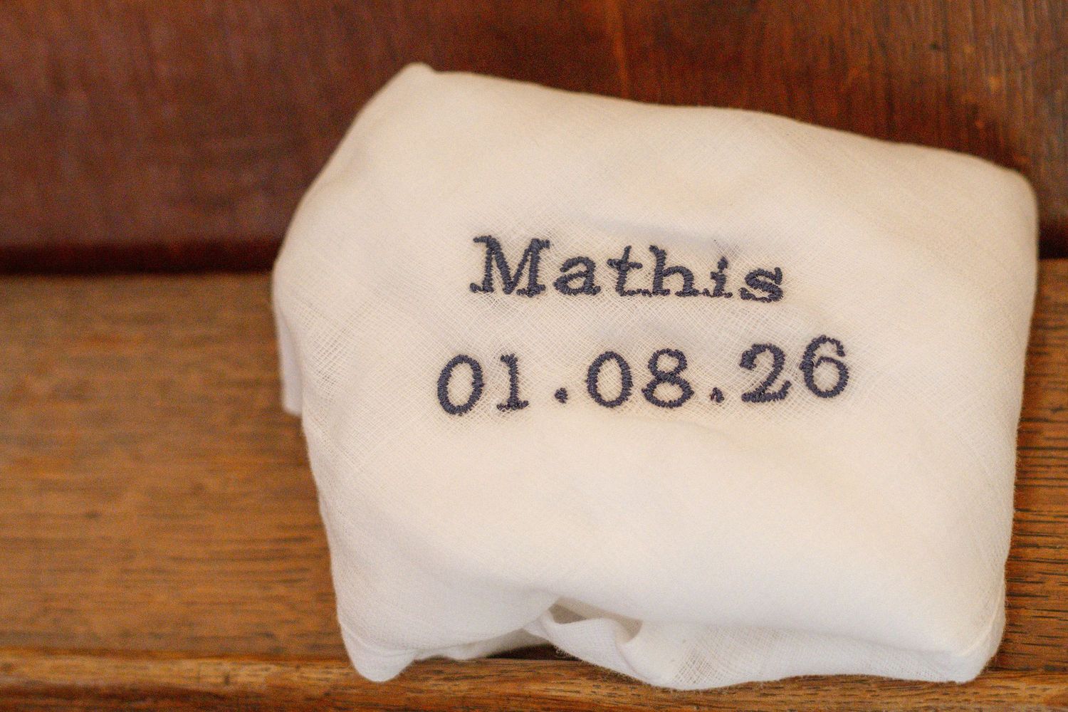 Souvenir en tissu blanc avec le nom brodé Mathis et la date 01.08.26, posé sur une surface en bois.