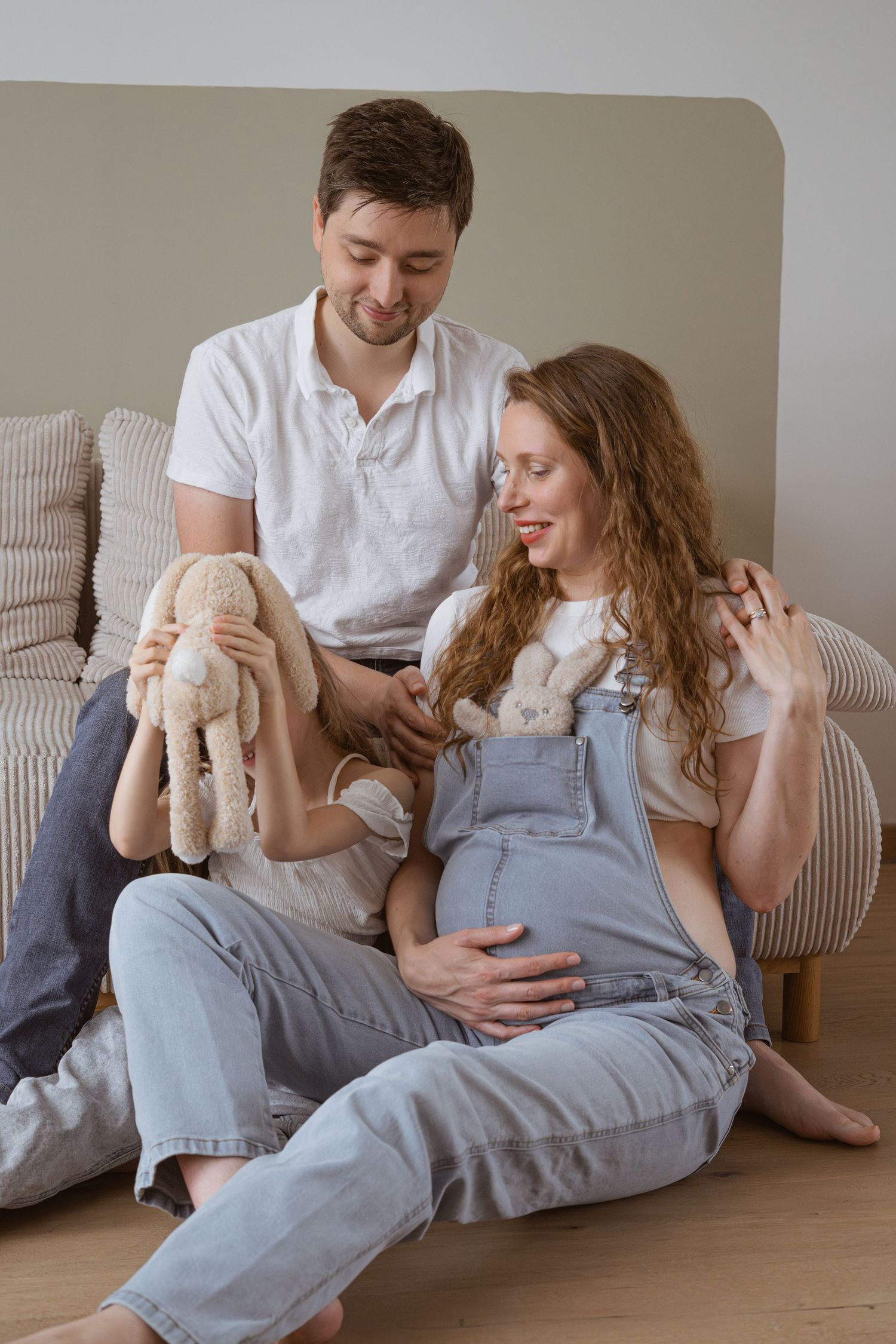 Happy pregnant couple sitting on floor with child holding stuffed bunny toy in cozy home setting.