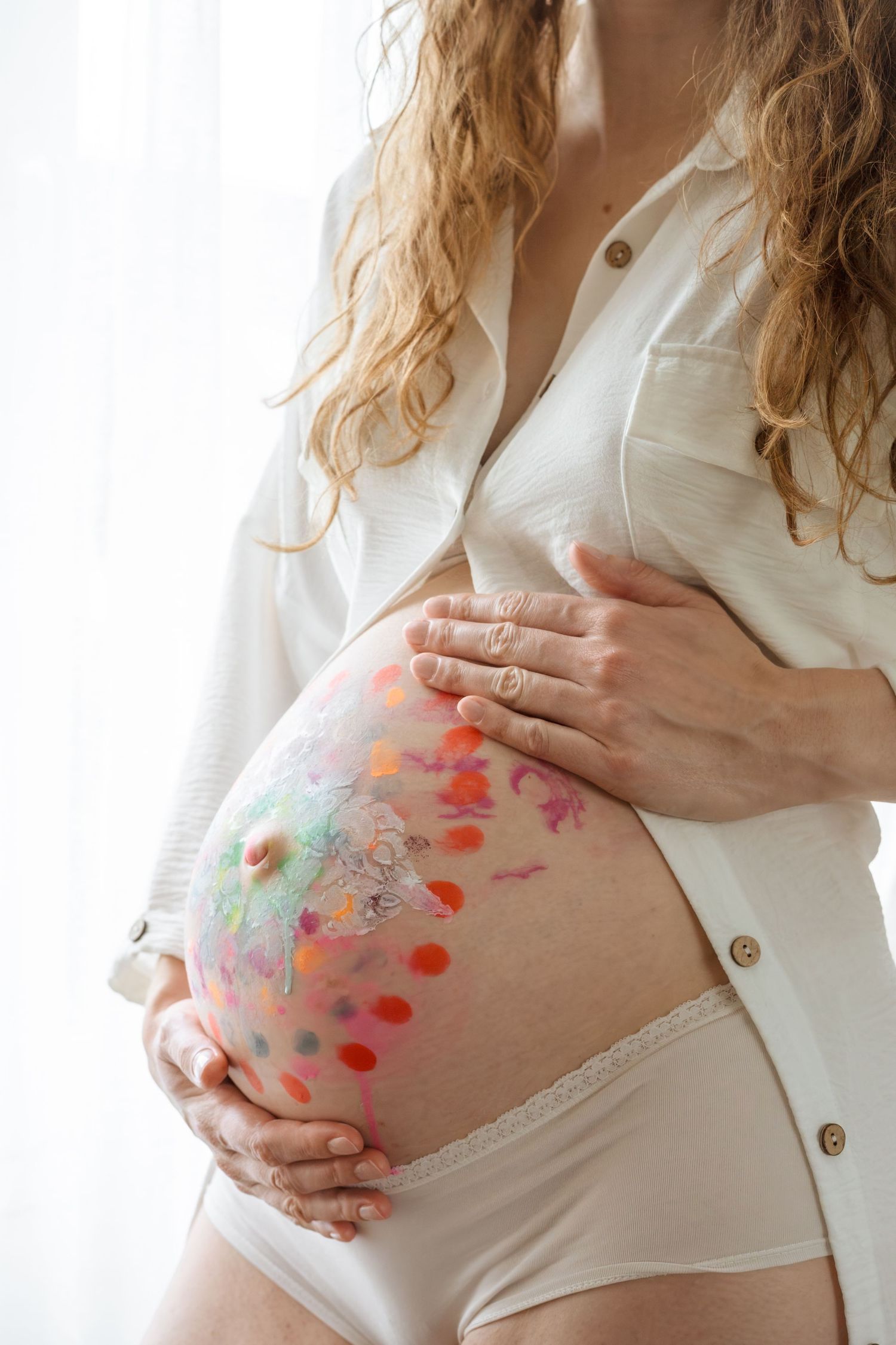 Pregnant woman in white shirt holding painted belly with colorful dots and designs near a bright window.