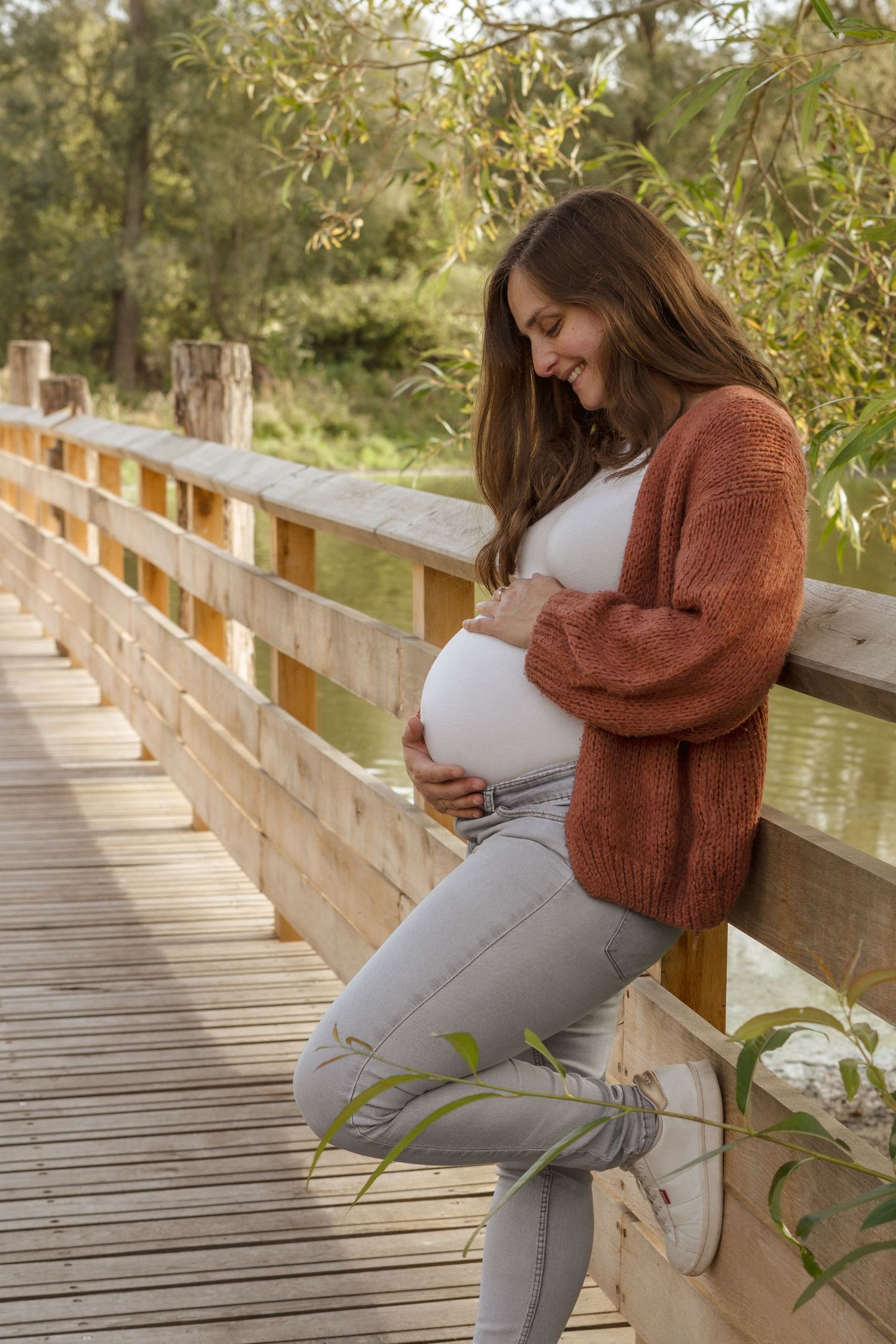 Pregnant woman in rust cardigan cradling baby bump on wooden bridge surrounded by lush greenery.