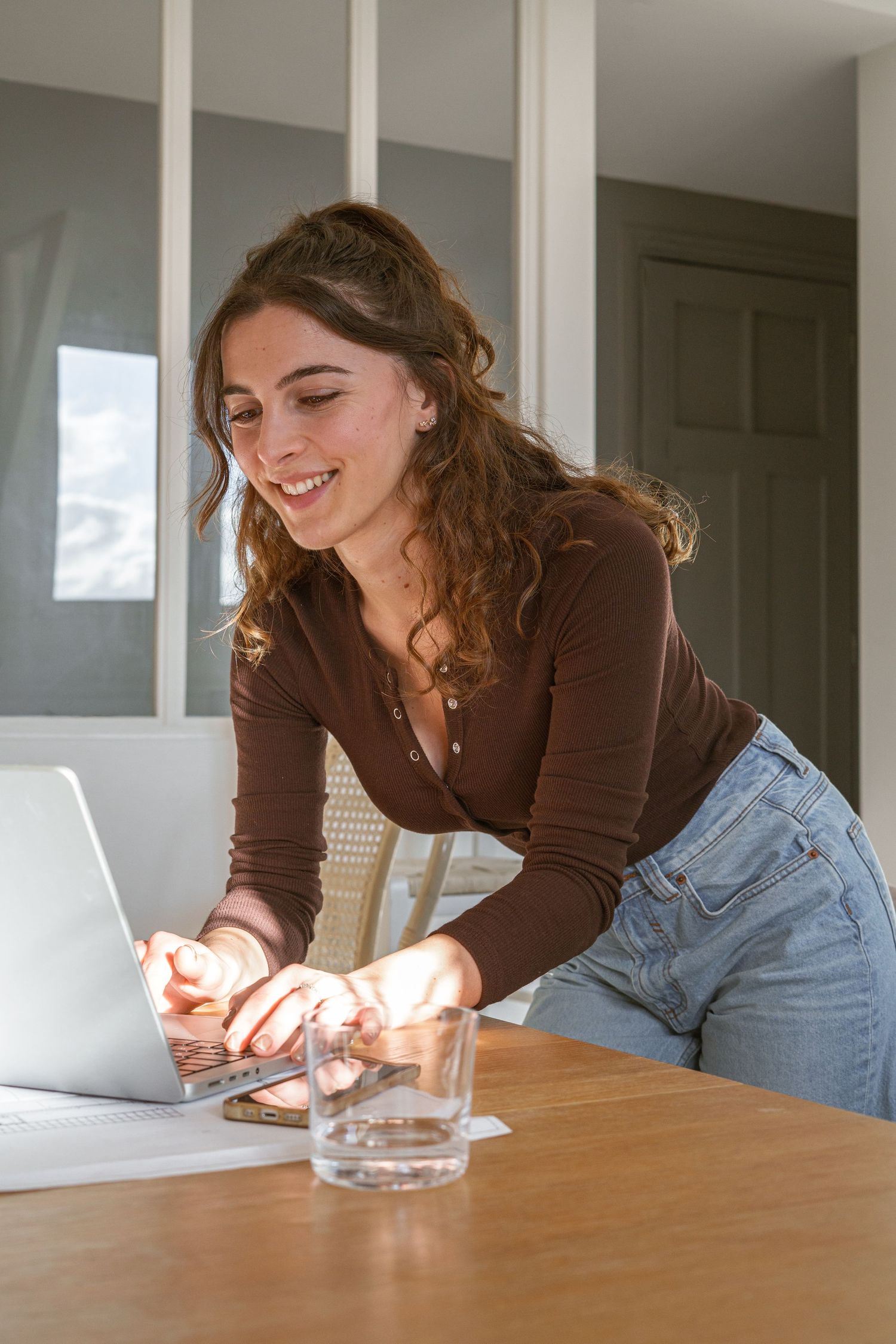 Smiling woman with wavy brown hair in a brown top and jeans leans over a wooden table typing on a laptop beside a glass of water.