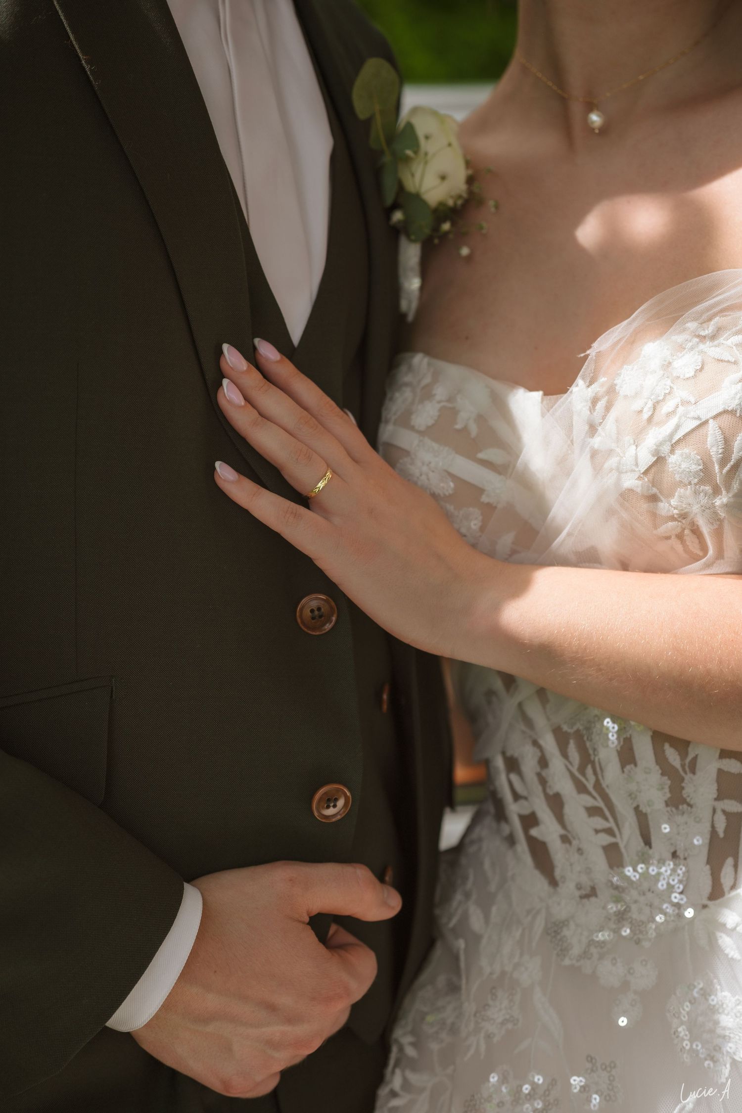 La mariée, vêtue d'une robe de mariée en dentelle et portant une alliance en or, pose sa main sur la veste de costume sombre du marié.