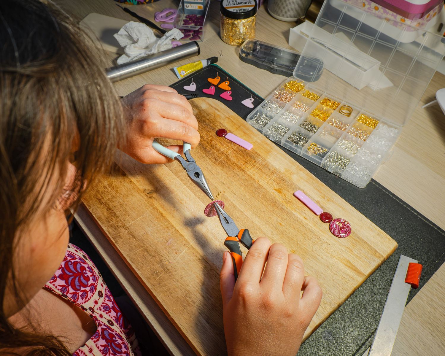 Young girl crafting jewelry at a wooden workbench with pliers, beads, and organizer trays in a creative workspace.