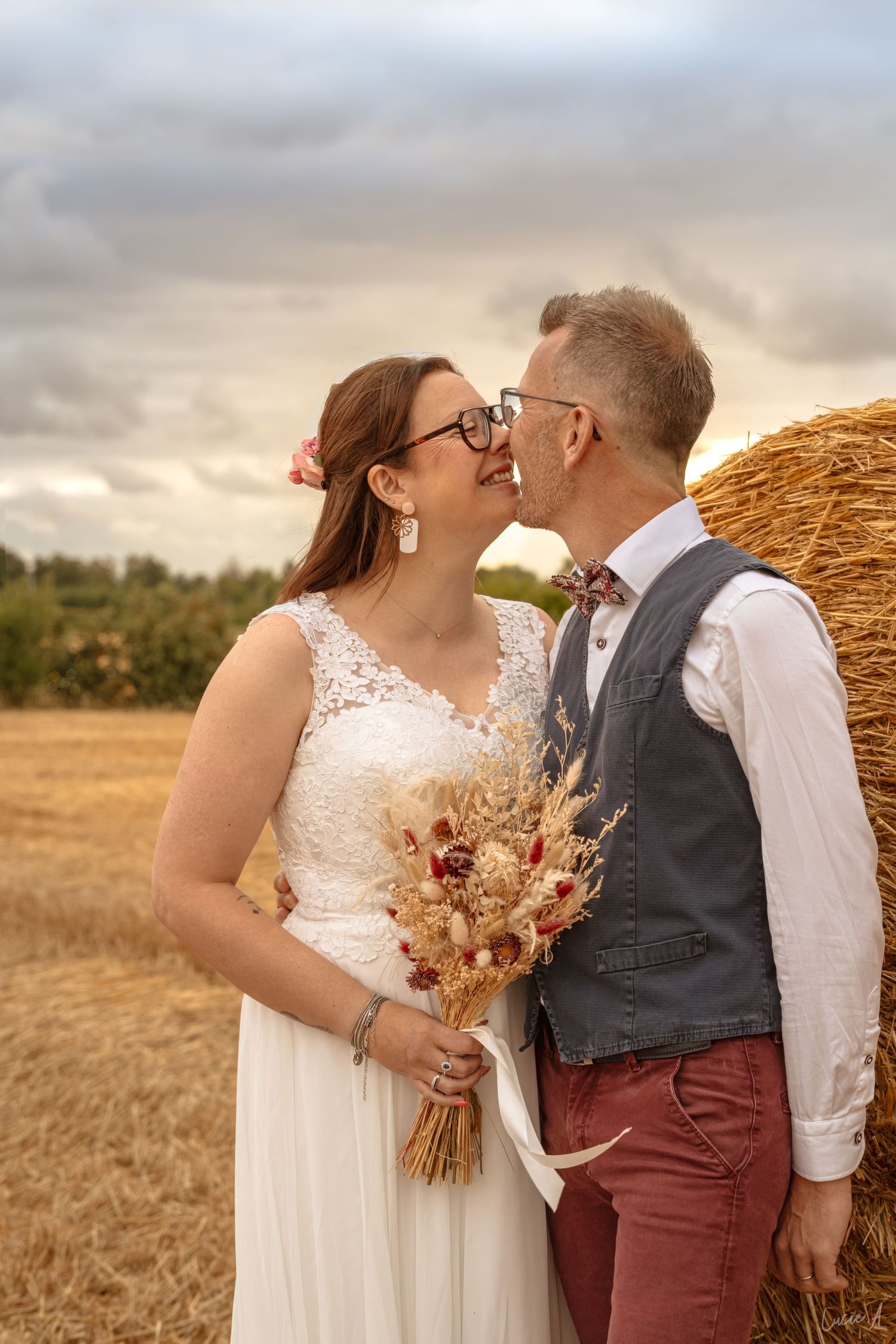 Dans un champ doré, les mariés échangent un baiser romantique près d'une botte de foin, la mariée tenant un bouquet de fleurs séchées.