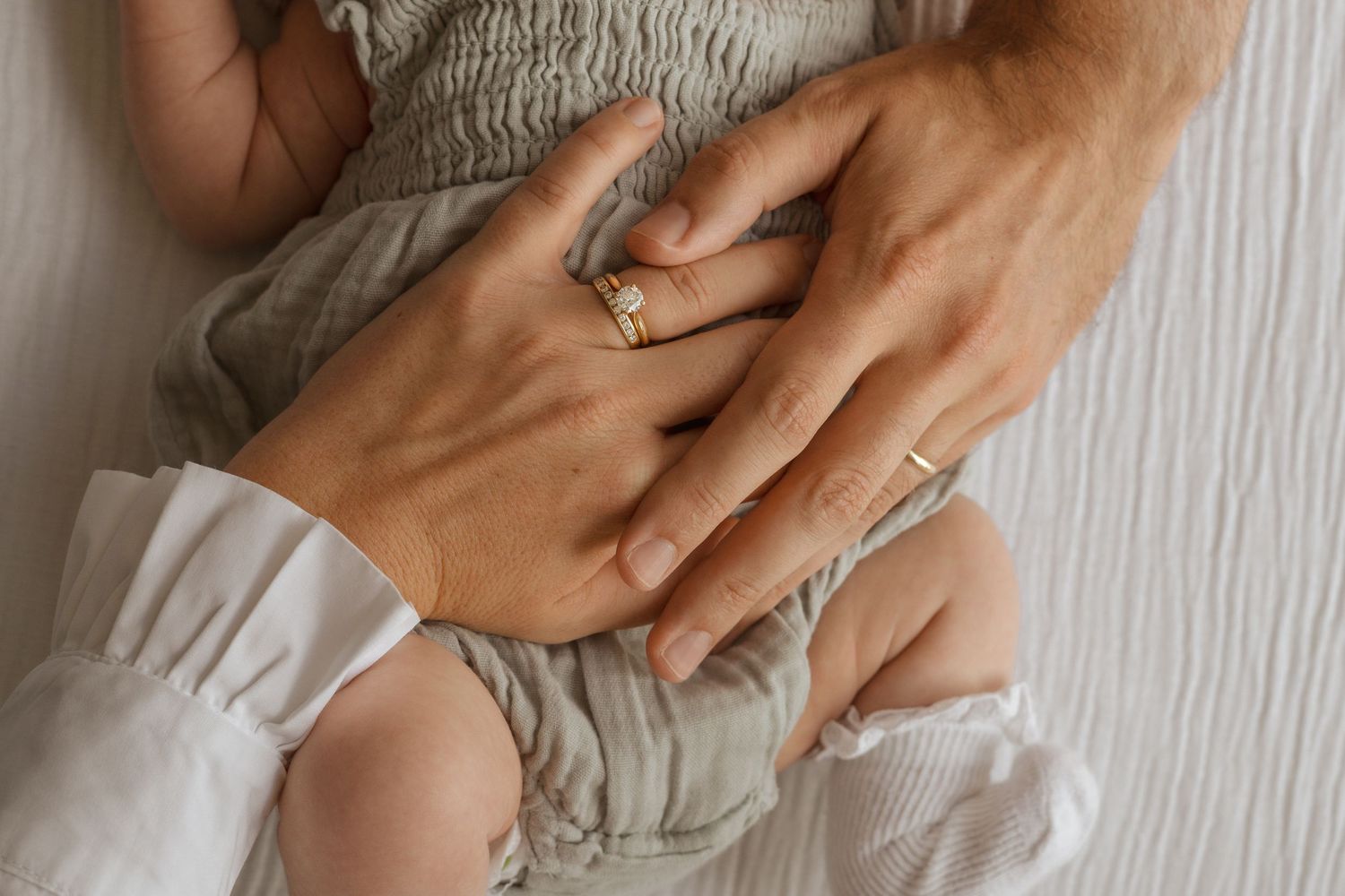 Two parents wearing gold wedding rings gently hold their newborn baby, capturing a tender family bonding moment.