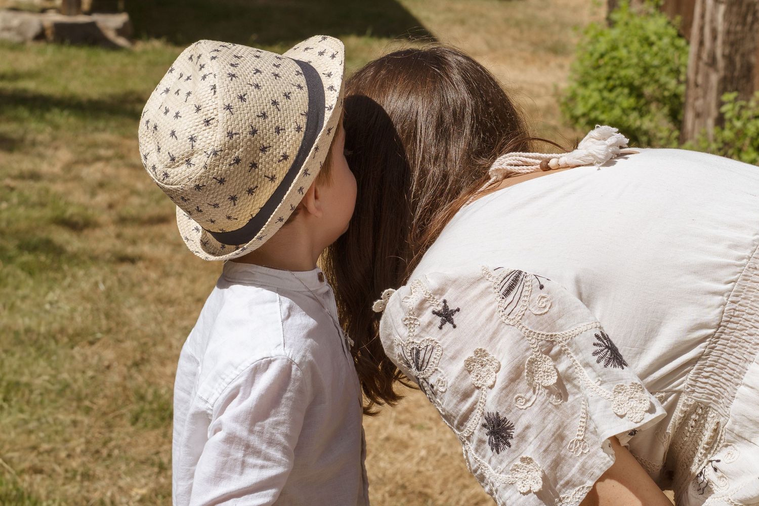 Un jeune enfant coiffé d'un chapeau de paille échange un tendre baiser avec une femme en robe de dentelle blanche, à l'extérieur.