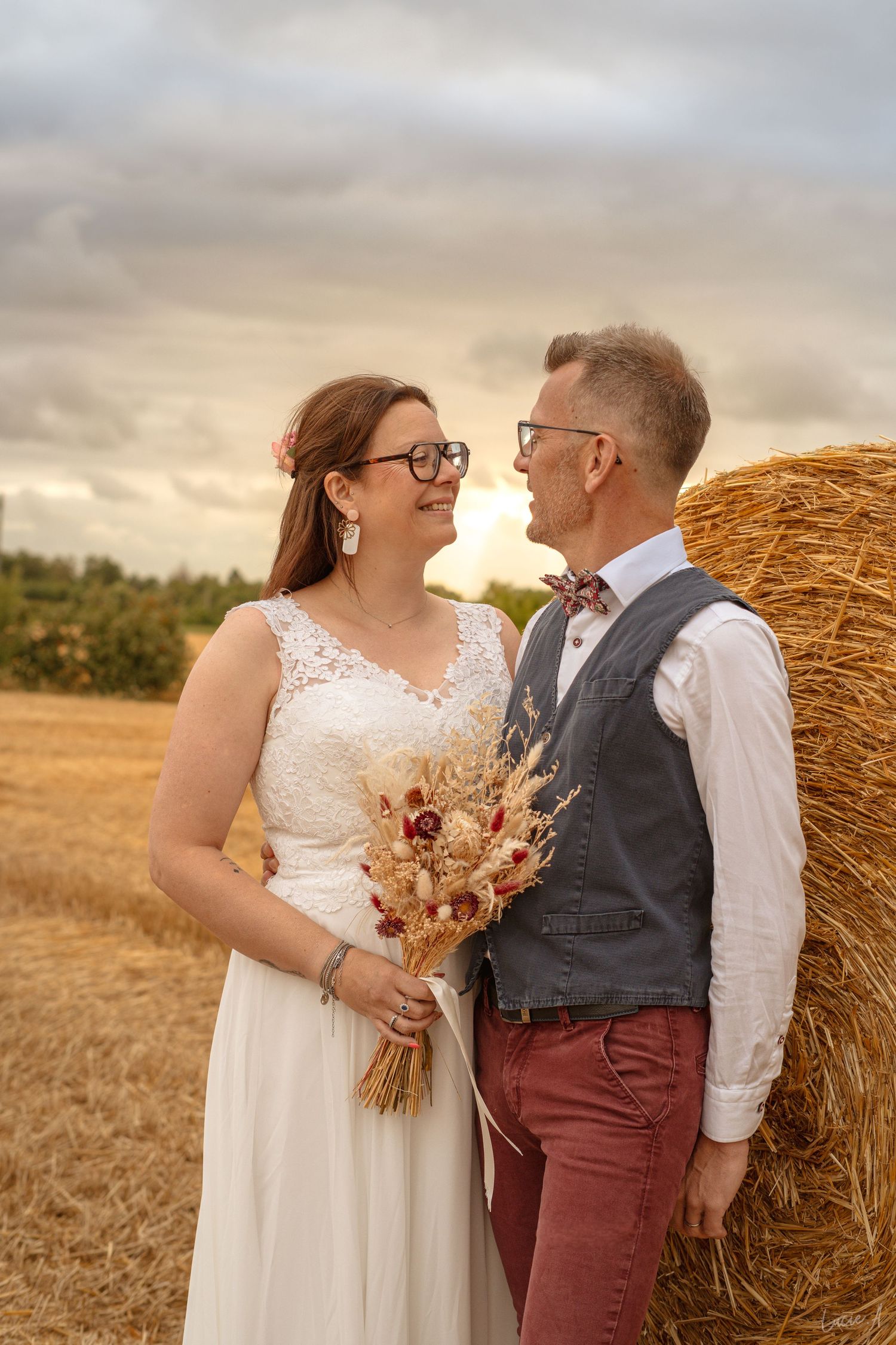Un couple de jeunes mariés portant des lunettes partage un moment romantique près d'une botte de foin dans un champ doré au coucher du solei