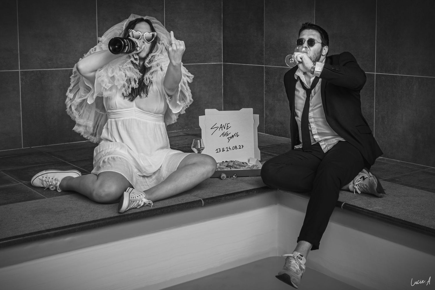Photo en noir et blanc de jeunes mariés assis sur un rebord, buvant à la bouteille, avec une boîte à pizza à côté d'eux.Séance save the date