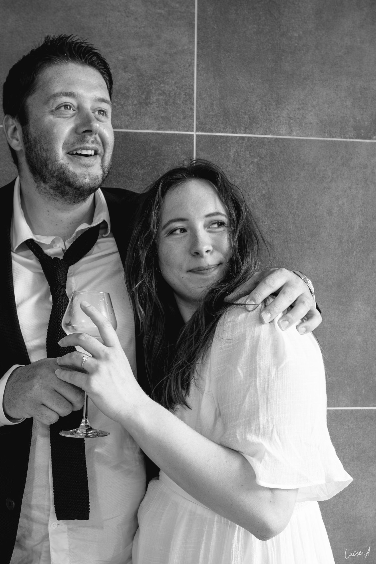 Photo en noir et blanc d'un couple souriant, l'homme en cravate tenant un verre de vin et le bras autour de la femme en robe blanche.