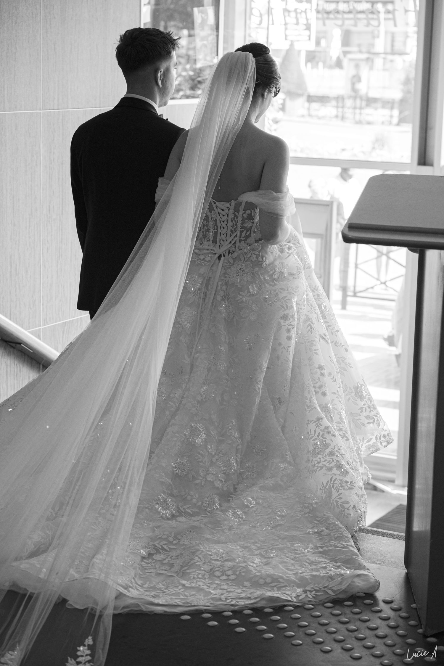 Photo en noir et blanc des mariés, vue de dos, mettant en valeur une robe de mariée en dentelle finement travaillée et un long voile fluide.