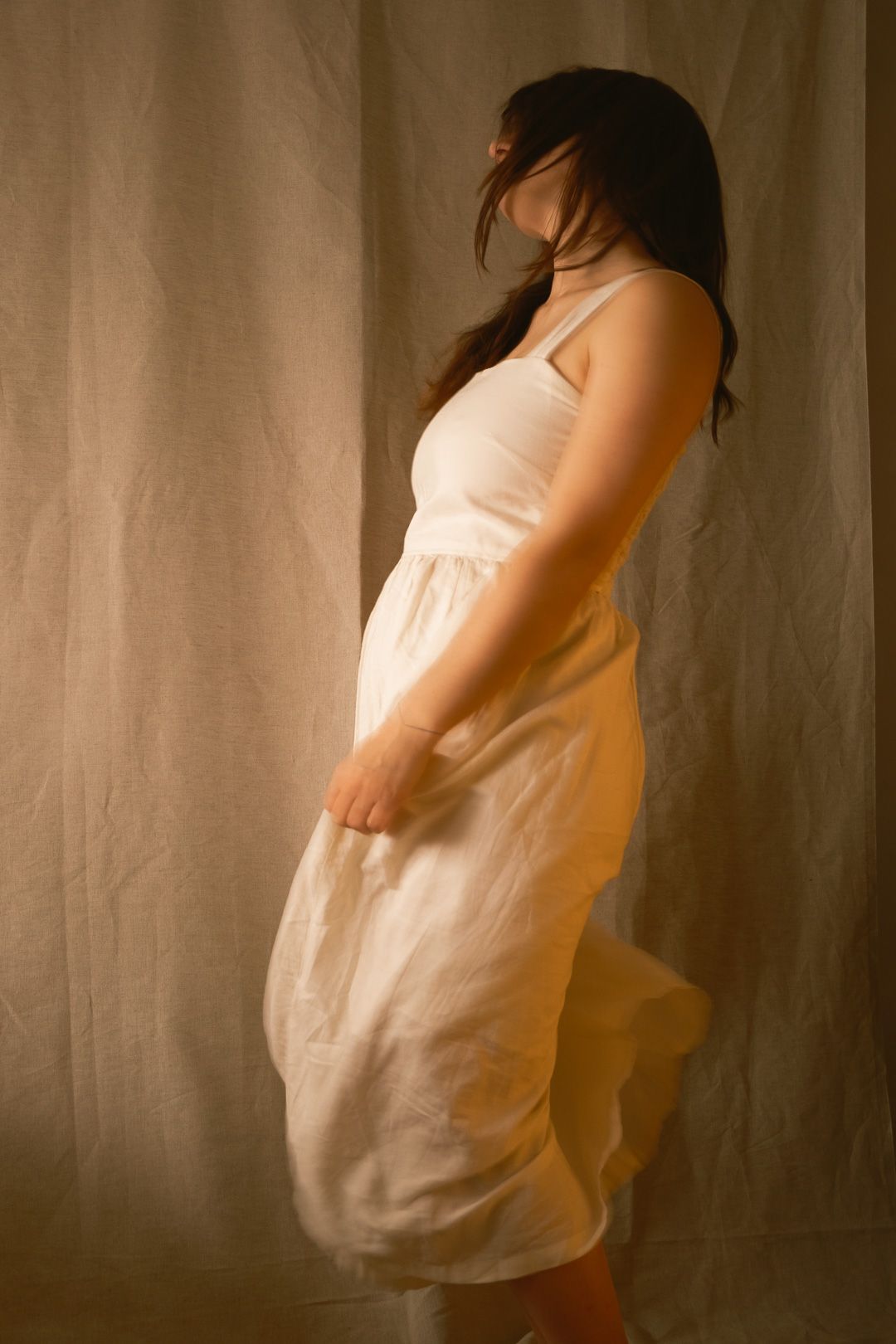 Femme en robe midi blanche fluide, debout de profil sur un fond de lin neutre dans une lumière de studio chaleureuse.
