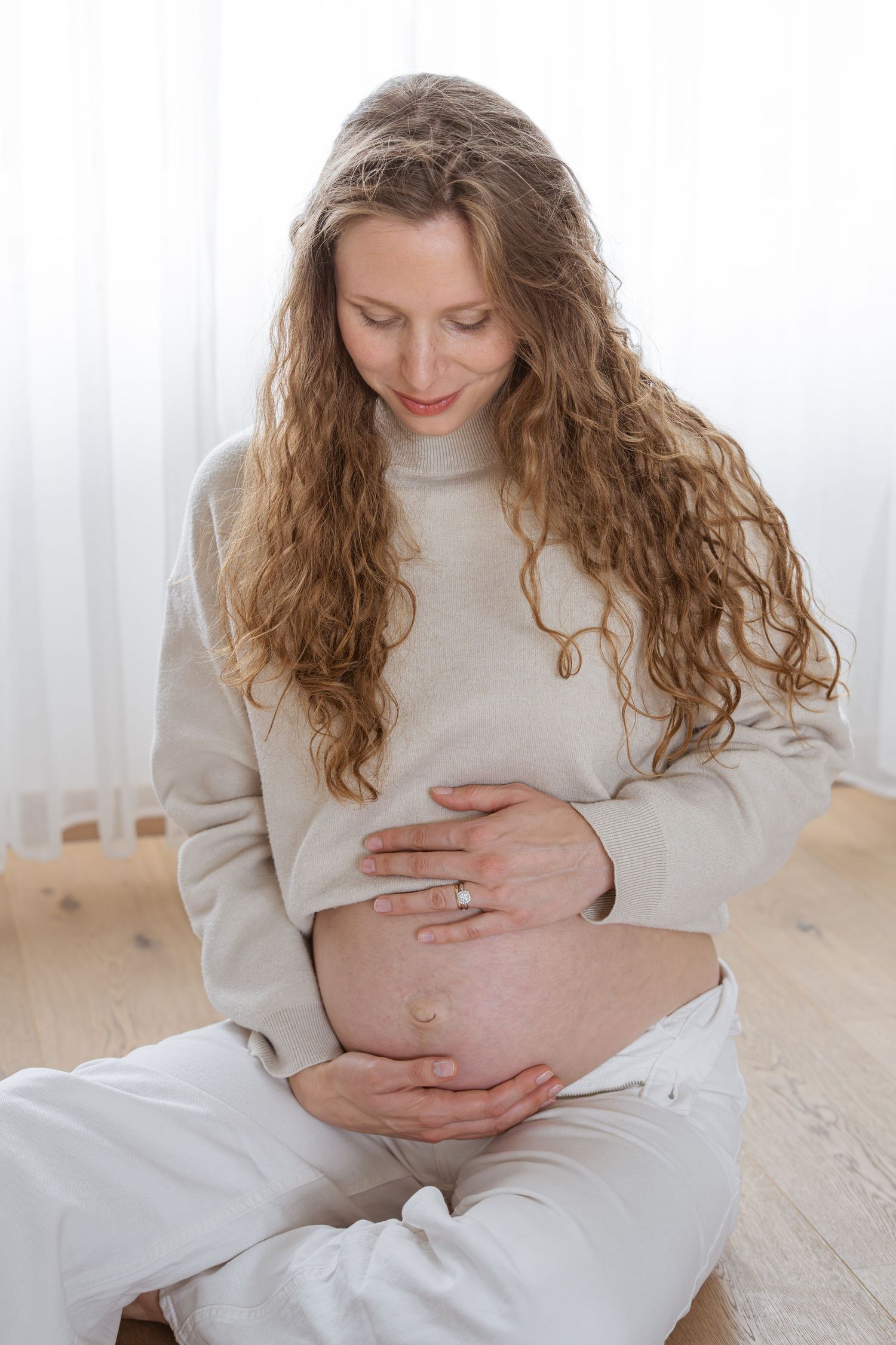 Pregnant woman with long curly hair cradling her bare baby bump while sitting on a wooden floor.
