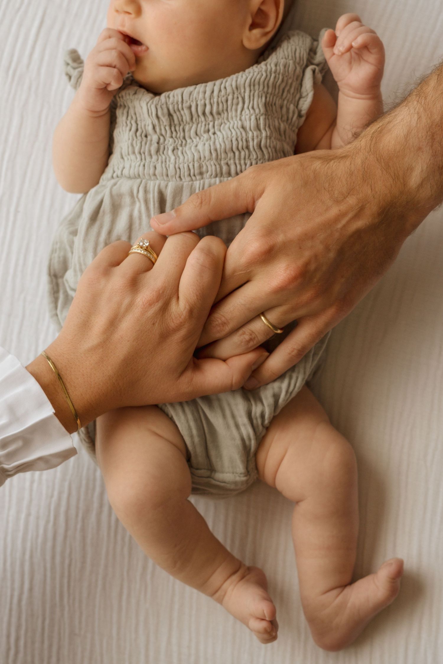 Parents' hands cradling a newborn baby in a sage green smocked romper on a white surface.
