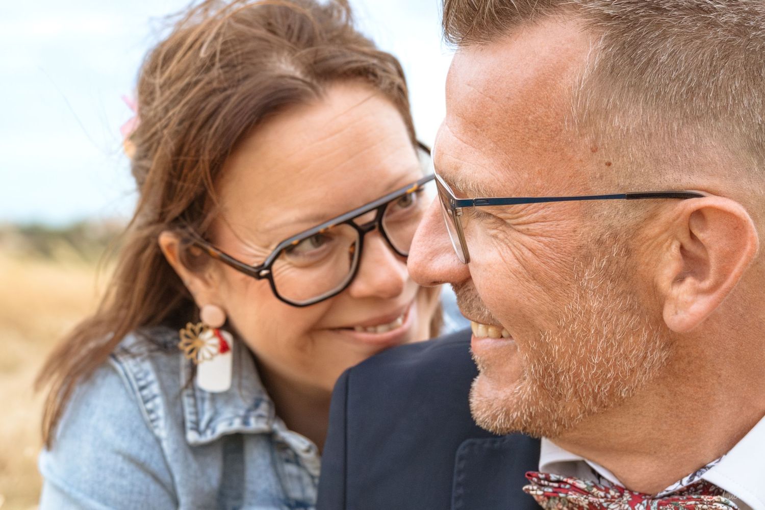Un couple souriant, portant des lunettes, partage un moment intime en plein air, dans un champ ensoleillé.