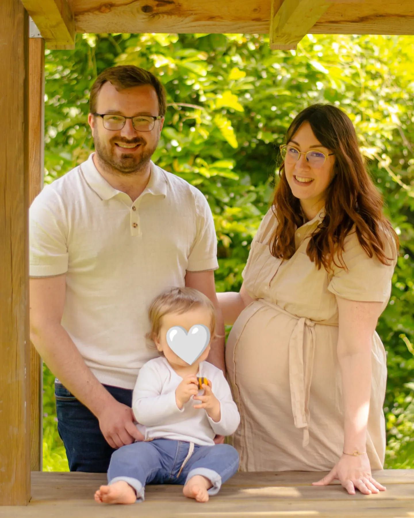 Une femme enceinte et un homme posent avec leur jeune enfant dans une tonnelle en bois entourée d'arbres verdoyants.