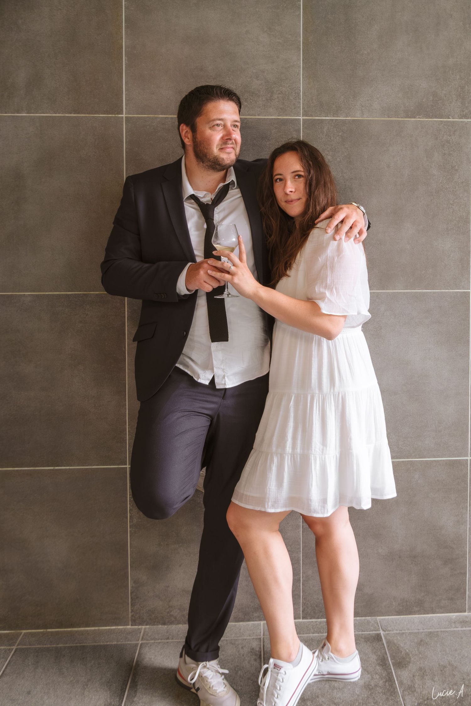 Un couple pose devant un mur carrelé gris, l'homme en costume et cravate, la femme en robe blanche et baskets.