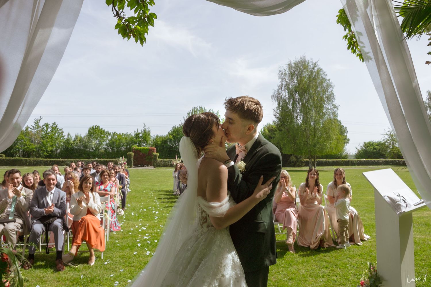 Un couple échange son premier baiser lors d'une cérémonie de mariage en plein air, les invités étant assis dans un jardin ensoleillé.