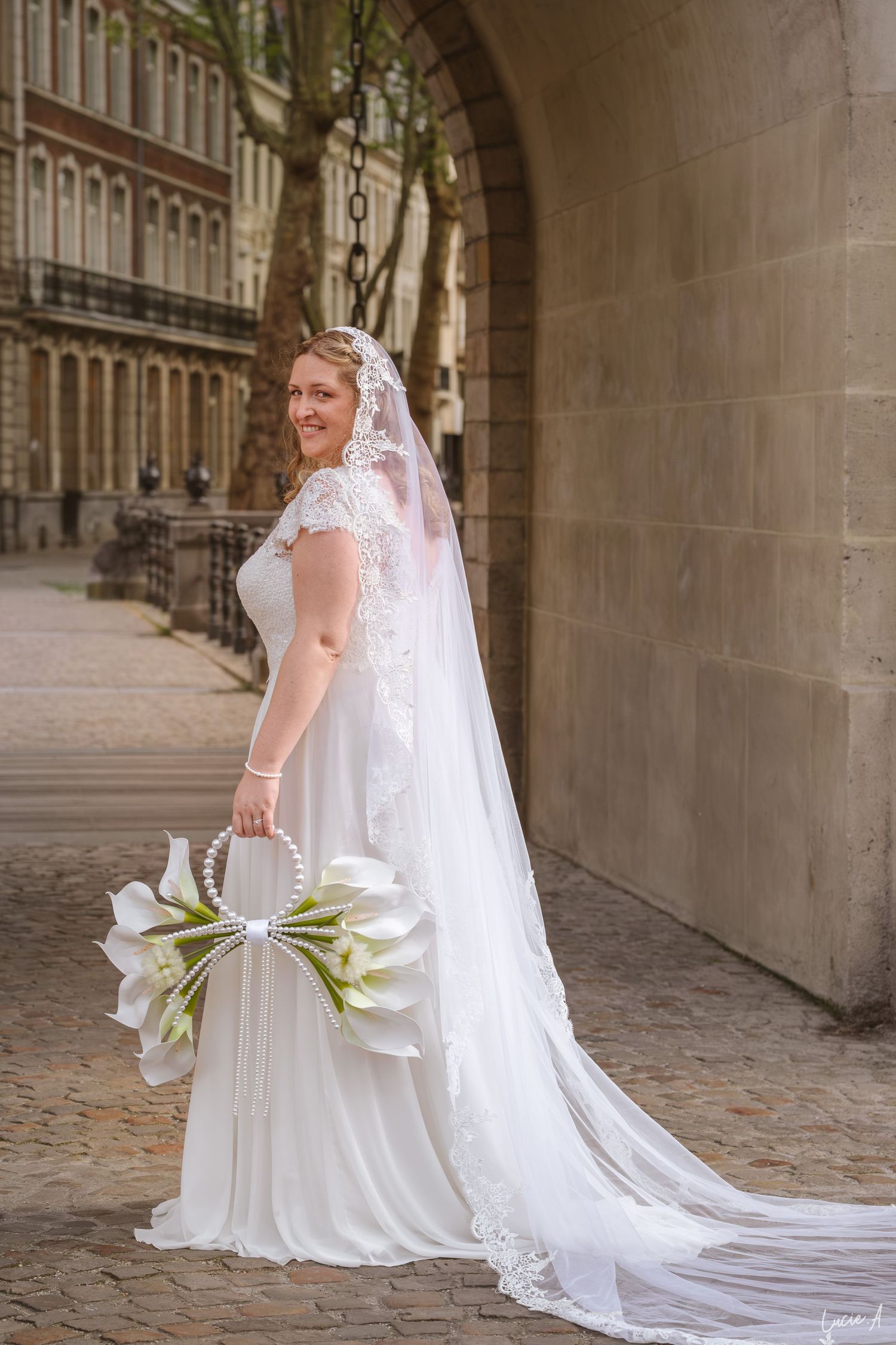 Une mariée en robe de dentelle blanche et voile tient un bouquet de lys calla dans une rue pavée près d'une arche en pierre.
