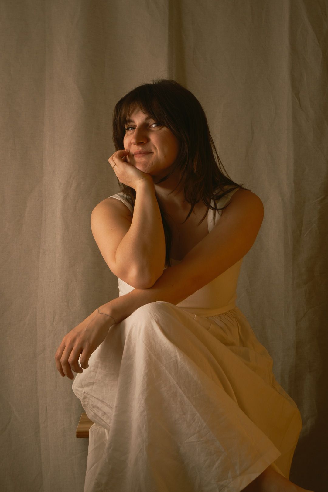 Une femme en robe blanche est assise avec grâce, le menton à la main, souriant doucement sur un fond neutre.
