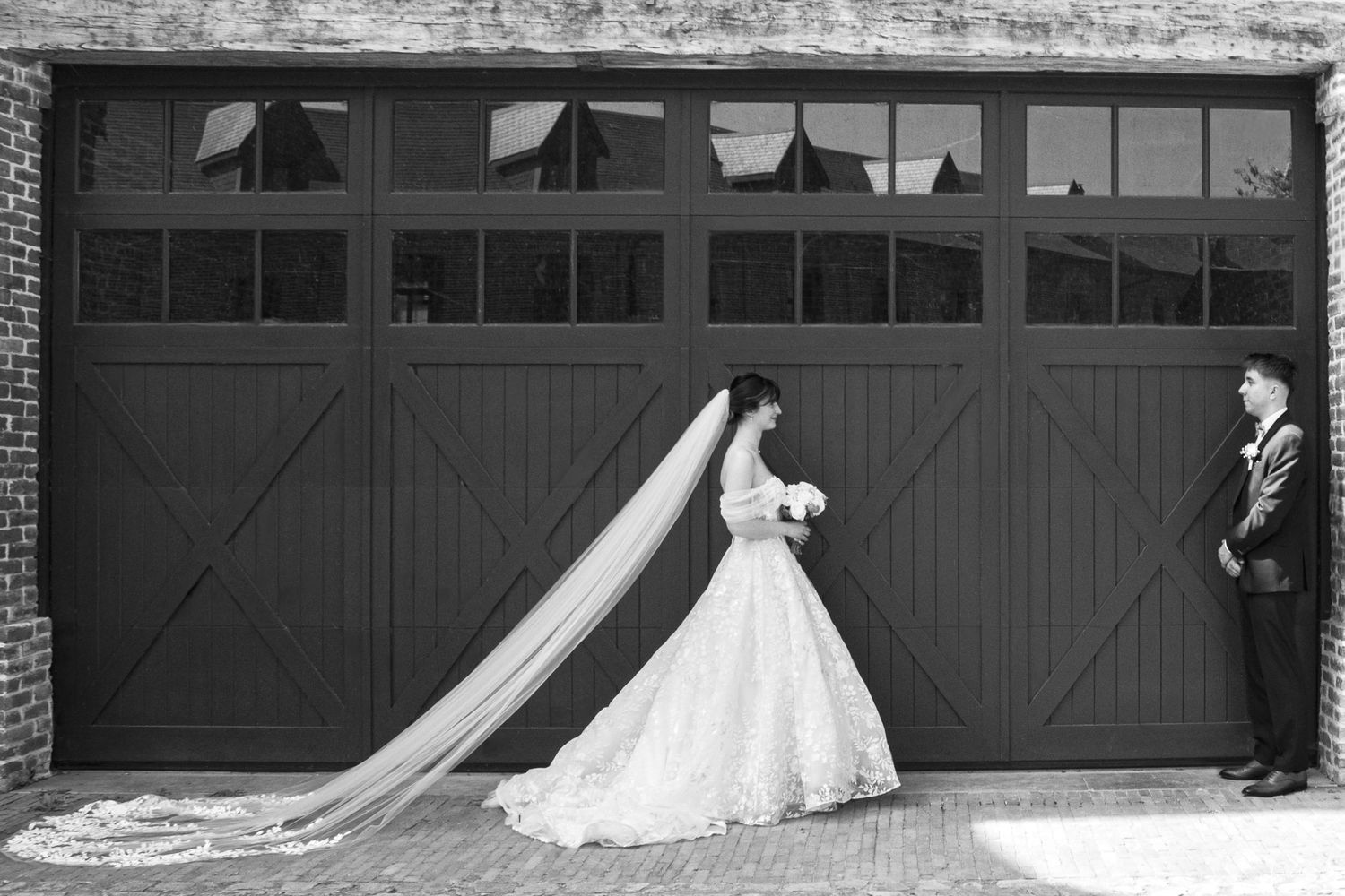 Photo en noir et blanc d'une mariée en robe à long voile et d'un marié debout devant de grandes portes de garage en bois.