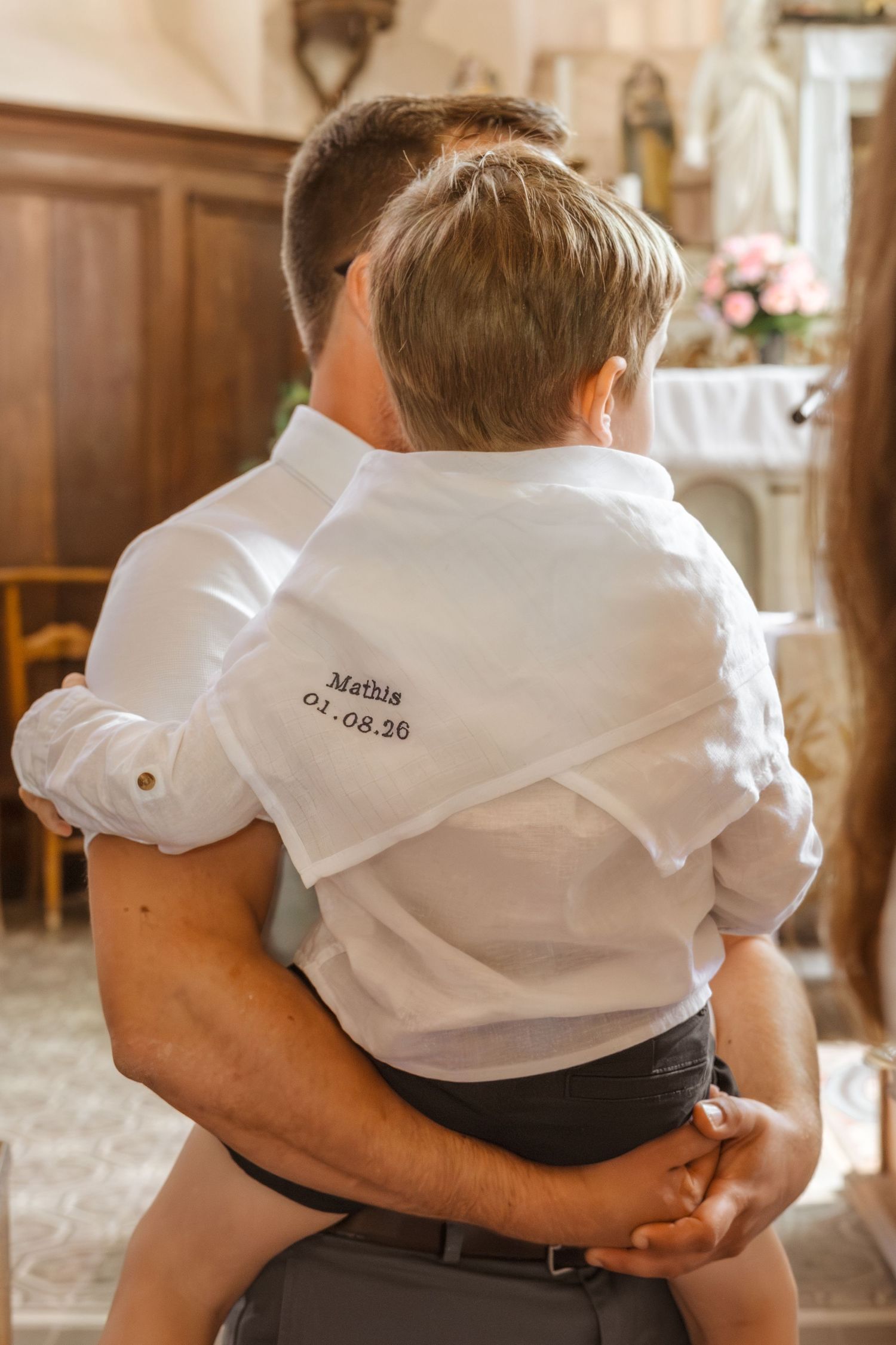 Adult holding child in white embroidered shirt reading Mathis 04.08.20 inside a church.