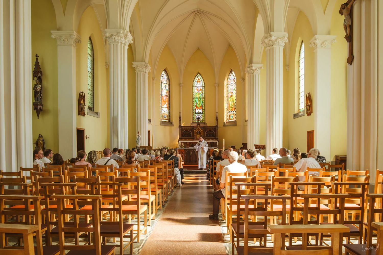 Interior of a Catholic church with wooden pews, white columns, stained glass windows, and congregation attending a religious service.