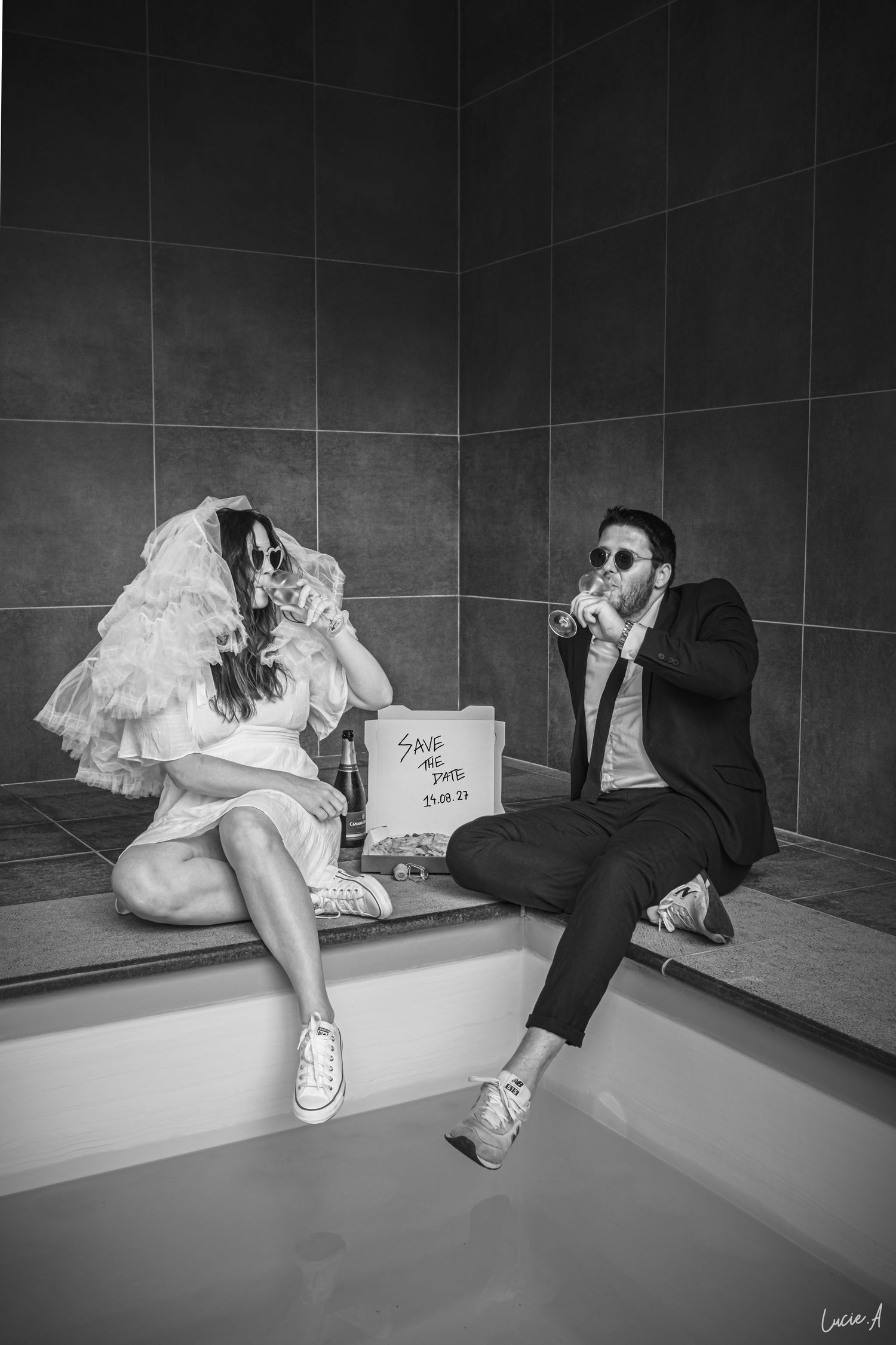 Photo en noir et blanc des mariés assis au bord d'une piscine, portant des lunettes de soleil et tenant des verres.