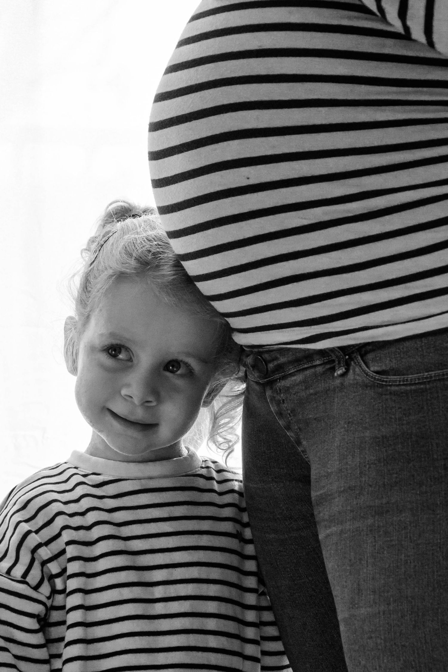 Portrait noir et blanc famille et grossesse, la petite fille se tient sous le ventre de sa mère en posant sa tête dessus, prise à Ronchin