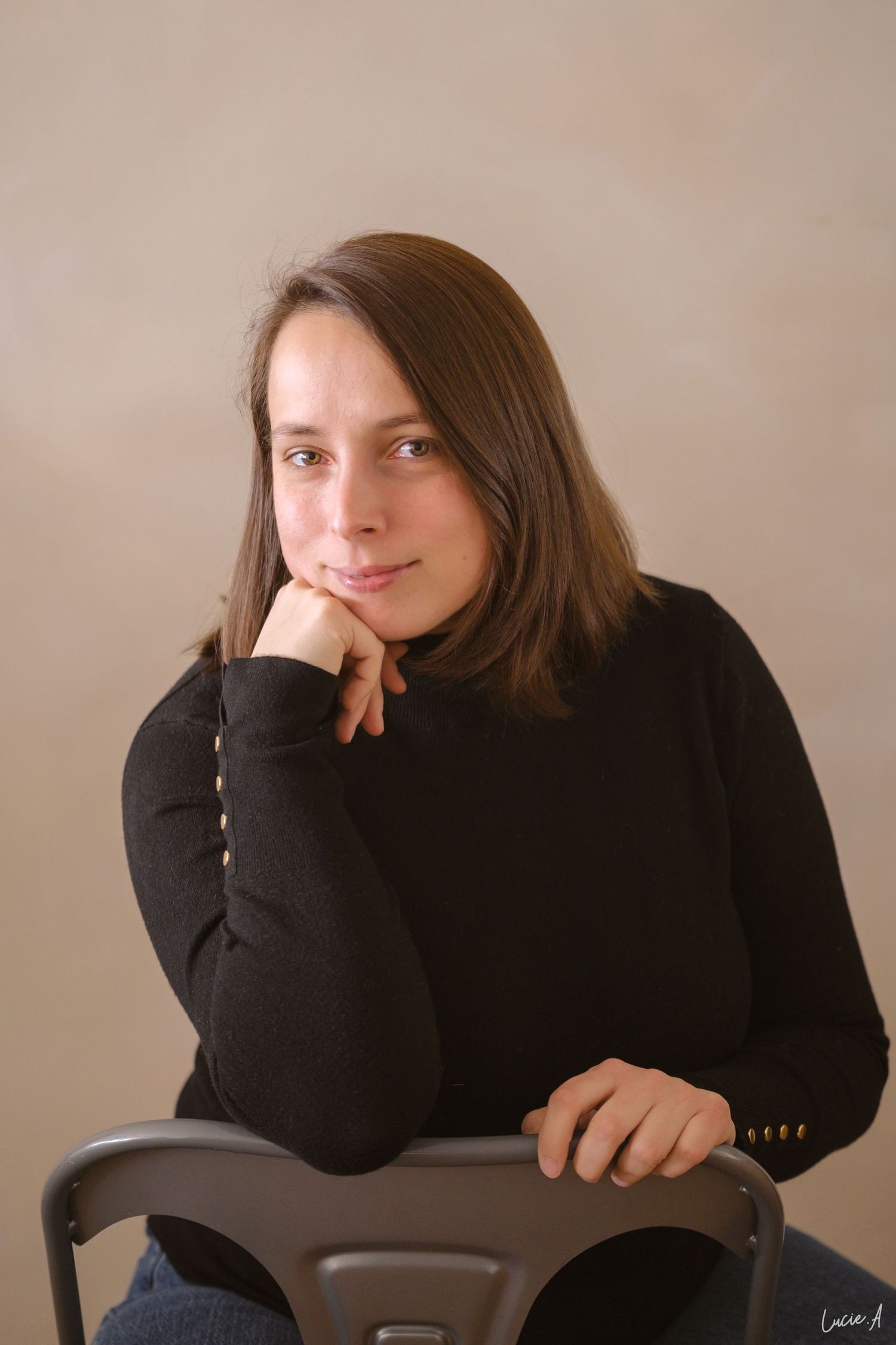 Jeune femme aux cheveux bruns, en col roulé noir, assise sur une chaise, le menton posé sur la main, souriant doucement. portrait corporate