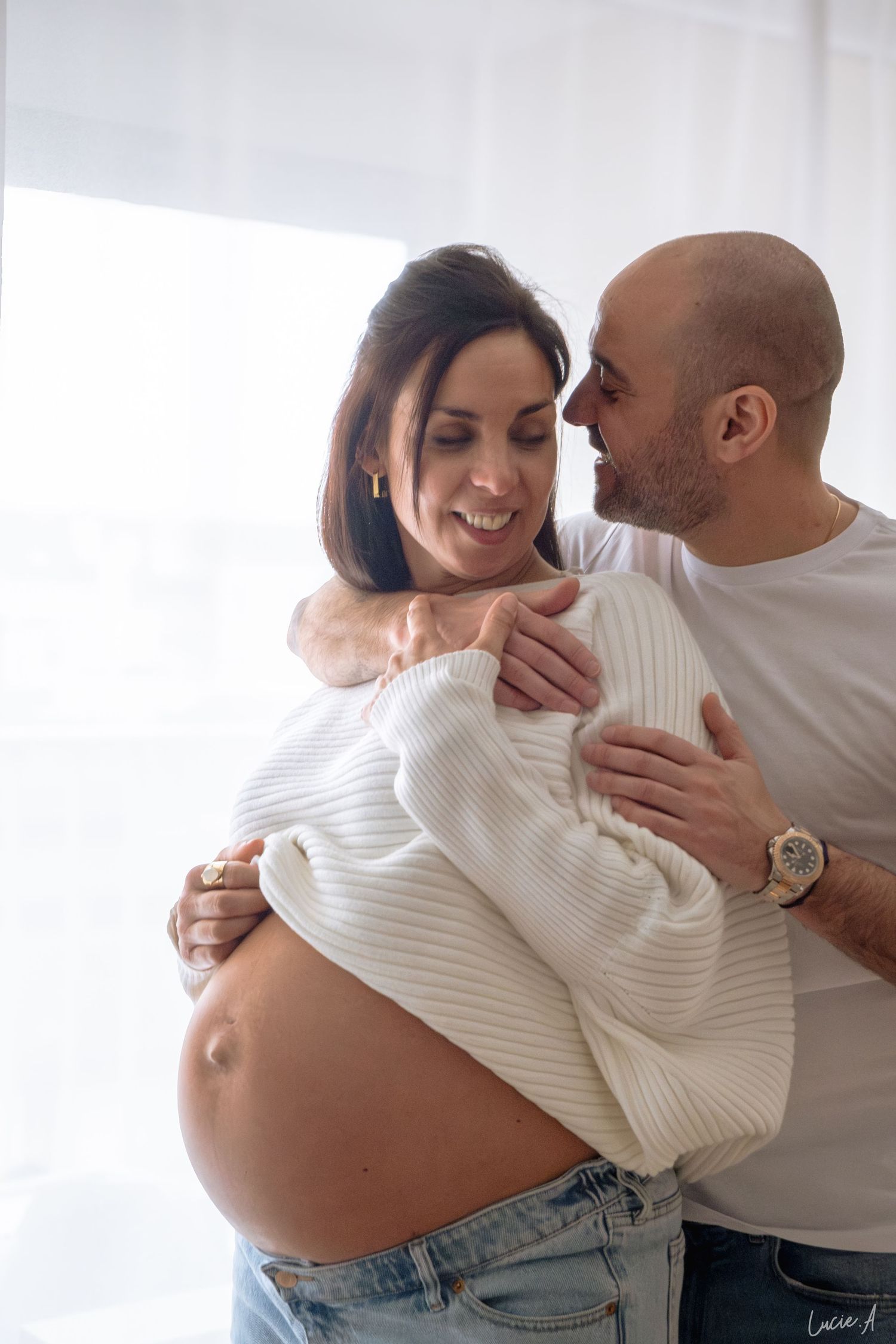 Une femme enceinte, radieuse, sourit, son partenaire l'enlaçant par derrière, dévoilant son ventre arrondi dans une pièce lumineuse.