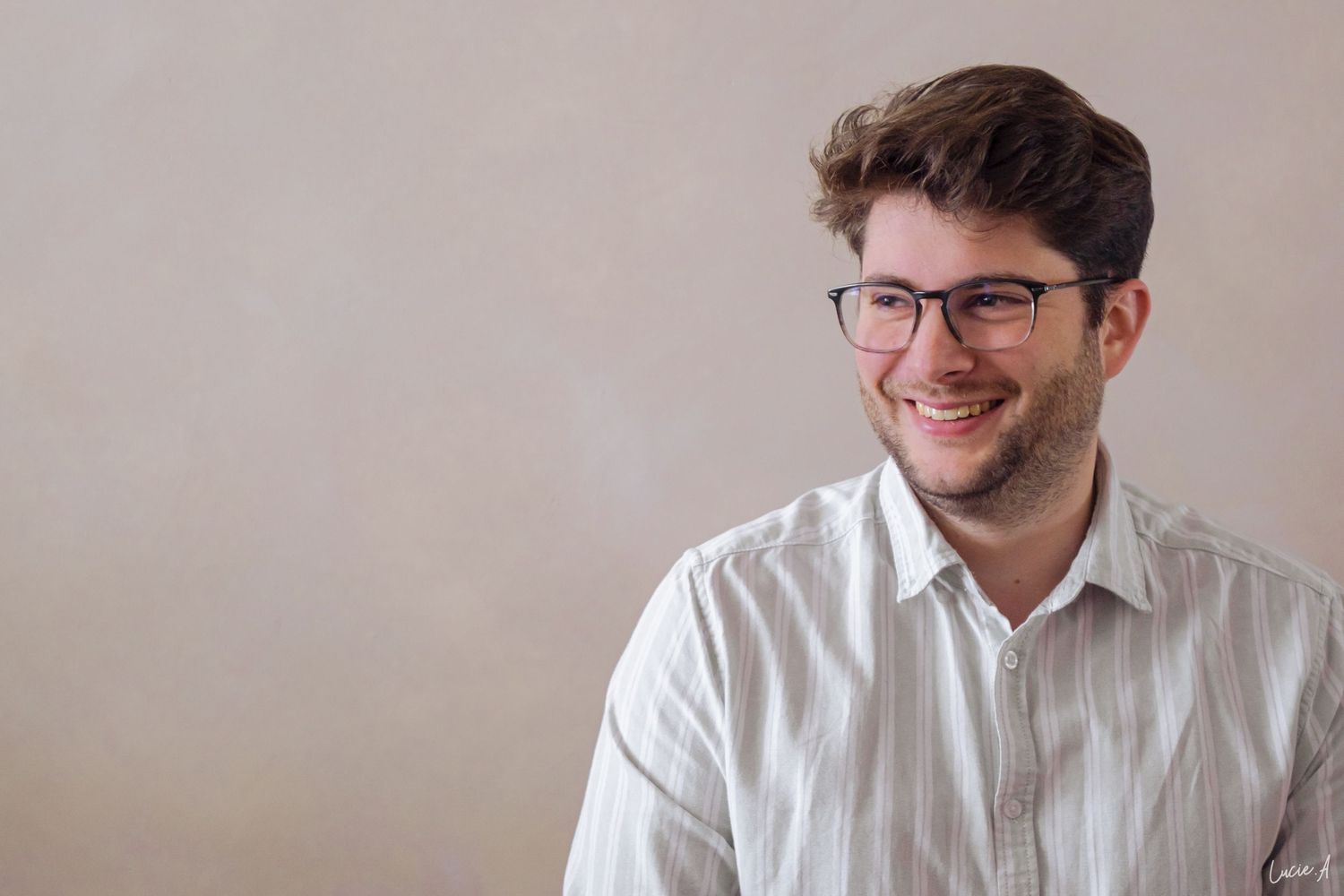 Smiling young man with glasses and curly hair wearing a striped shirt against a neutral background.