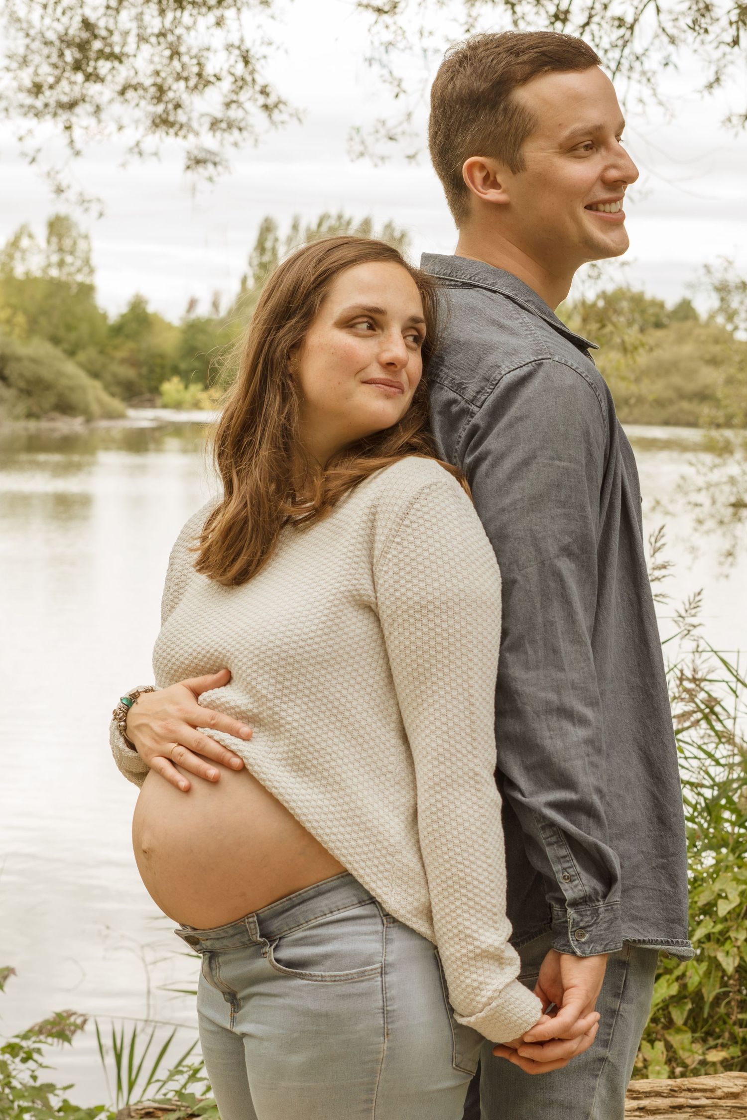 Pregnant couple standing back-to-back by a lake, woman cradling her baby bump, both smiling in a serene outdoor setting.