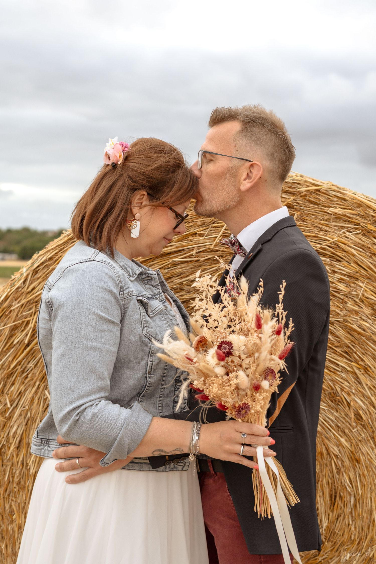 Newlywed couple sharing a romantic kiss in front of a hay bale, bride holding dried flower bouquet outdoors.