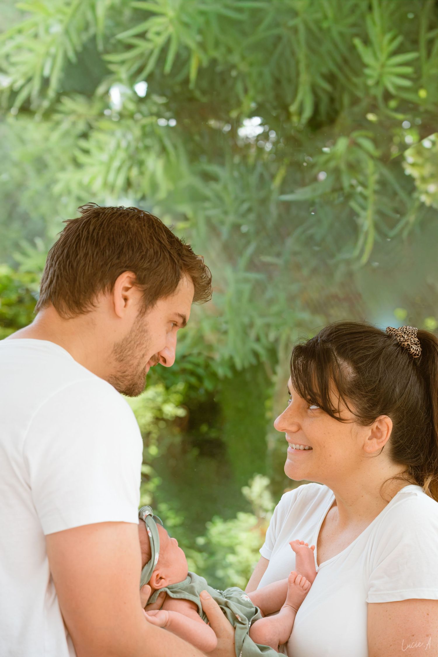 Un couple en chemises blanches sourit à son nouveau-né à l'extérieur, entouré d'arbres verdoyants.