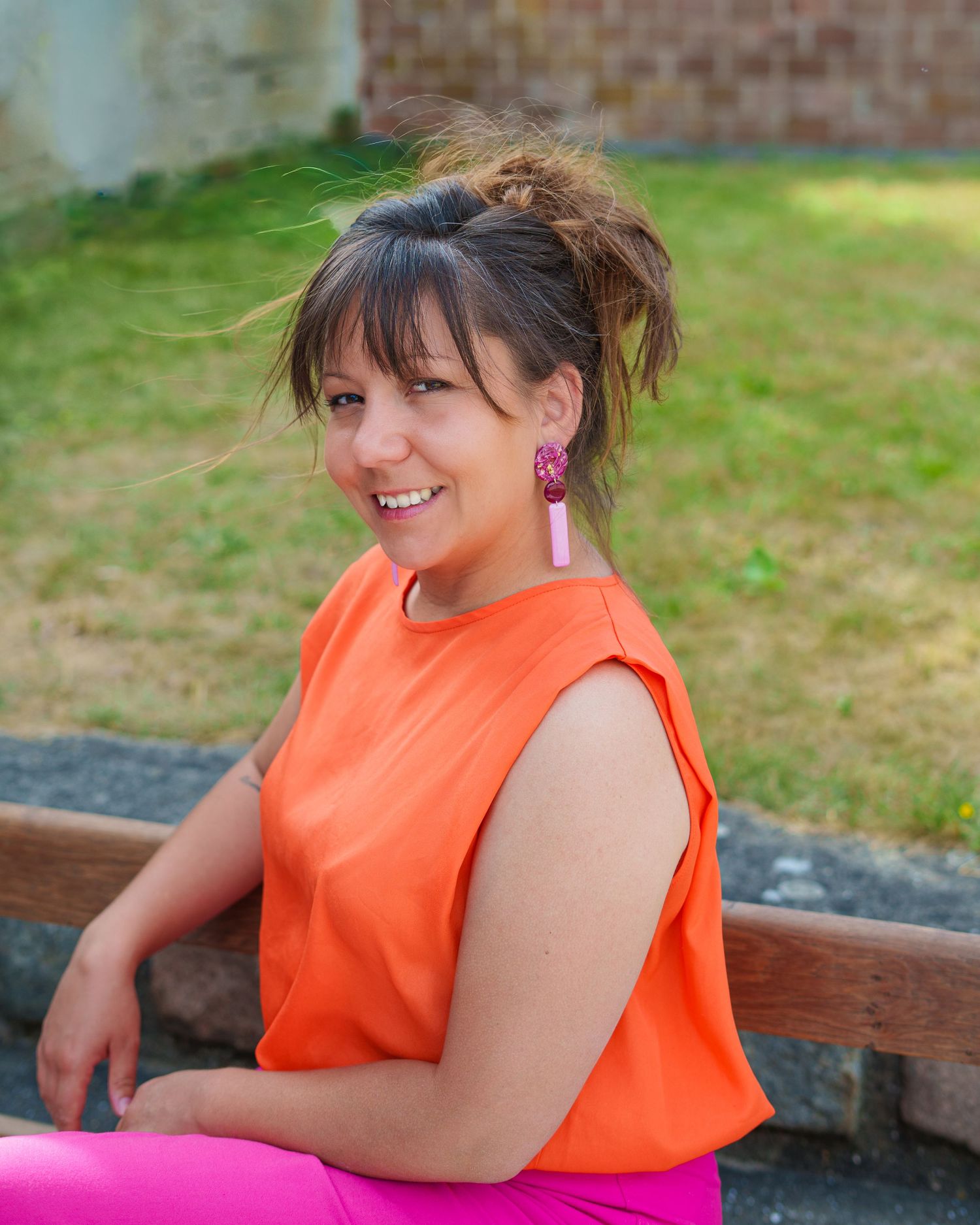 Jeune femme souriante, vêtue d'un débardeur orange et de boucles d'oreilles roses, assise sur un banc à l'extérieur.