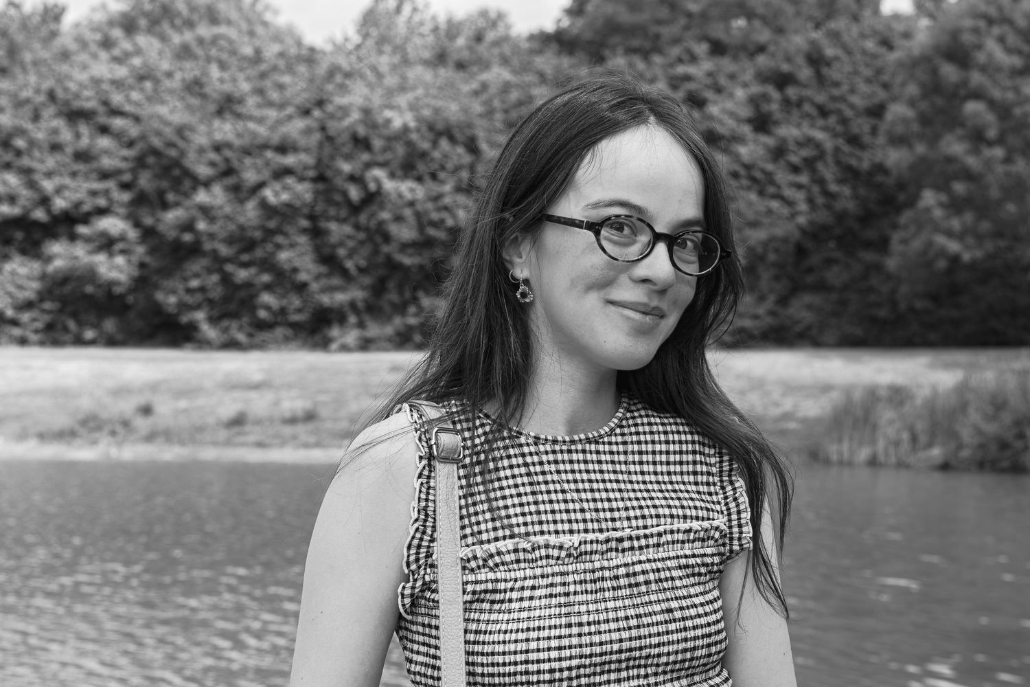 Jeune femme à lunettes et aux longs cheveux souriant en extérieur près d'un lac, sur une photo en noir et blanc.
