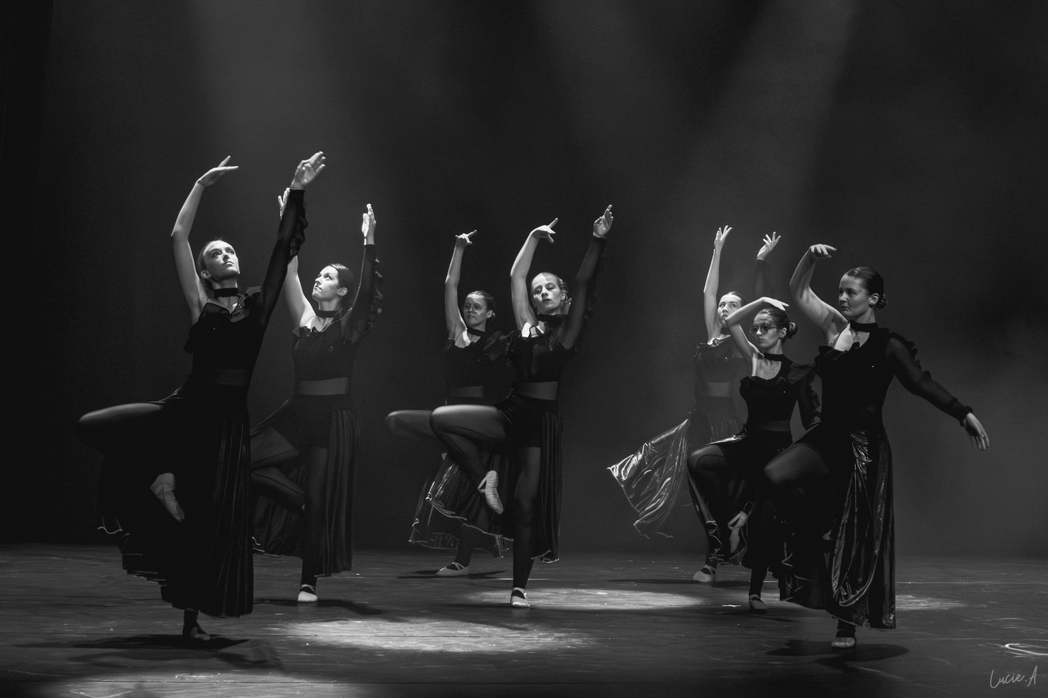 Photo en noir et blanc de danseurs en tenue noire se produisant sur une scène spectaculaire éclairée par des projecteurs.